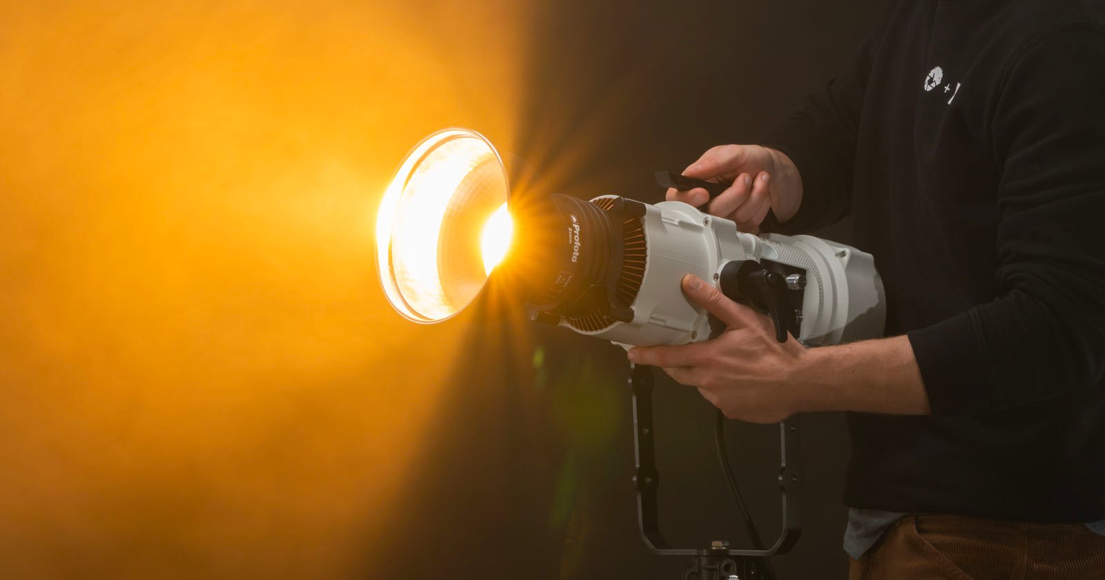 A person holding an LED light with a large attached reflector, aiming a bright light towards the left, in a studio with a warm, orange-hued backdrop.