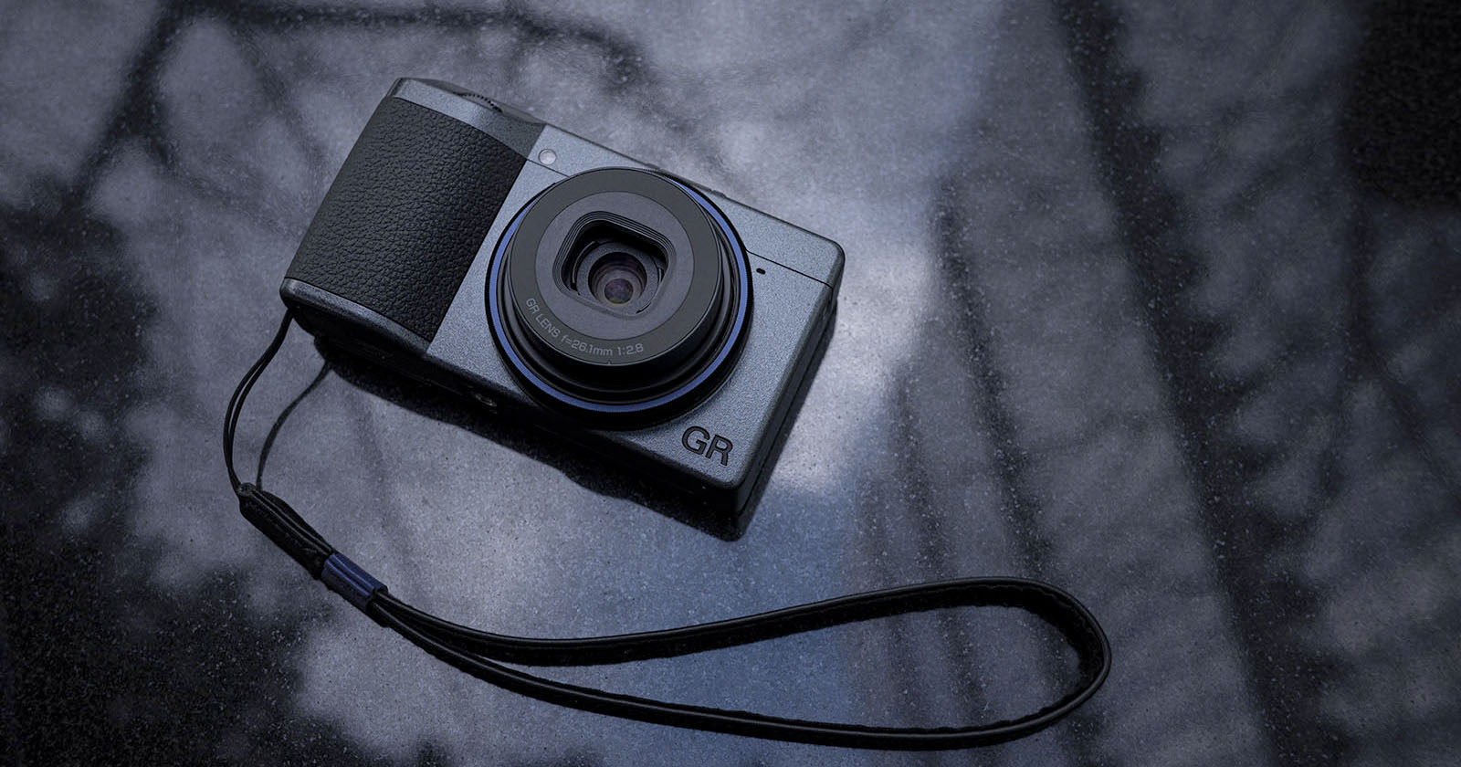 A compact digital camera with a black and gray design rests on a reflective surface. It features a visible lens and a wrist strap attached to the side. The surface has a blurred, abstract pattern in the background. A compact digital camera with a black and gray design rests on a reflective surface. It features a visible lens and a wrist strap attached to the side. The surface has a blurred, abstract pattern in the background.