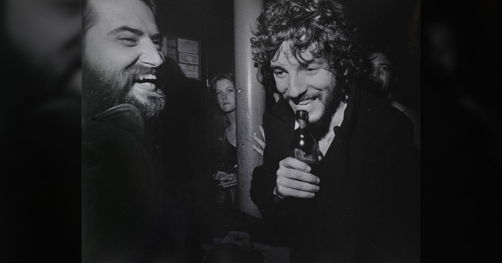 Two men laughing together at a bar, one holding a bottle, with a woman and another person in the background; black and white photo, candid and lively atmosphere. Two men laughing together at a bar, one holding a bottle, with a woman and another person in the background; black and white photo, candid and lively atmosphere.
