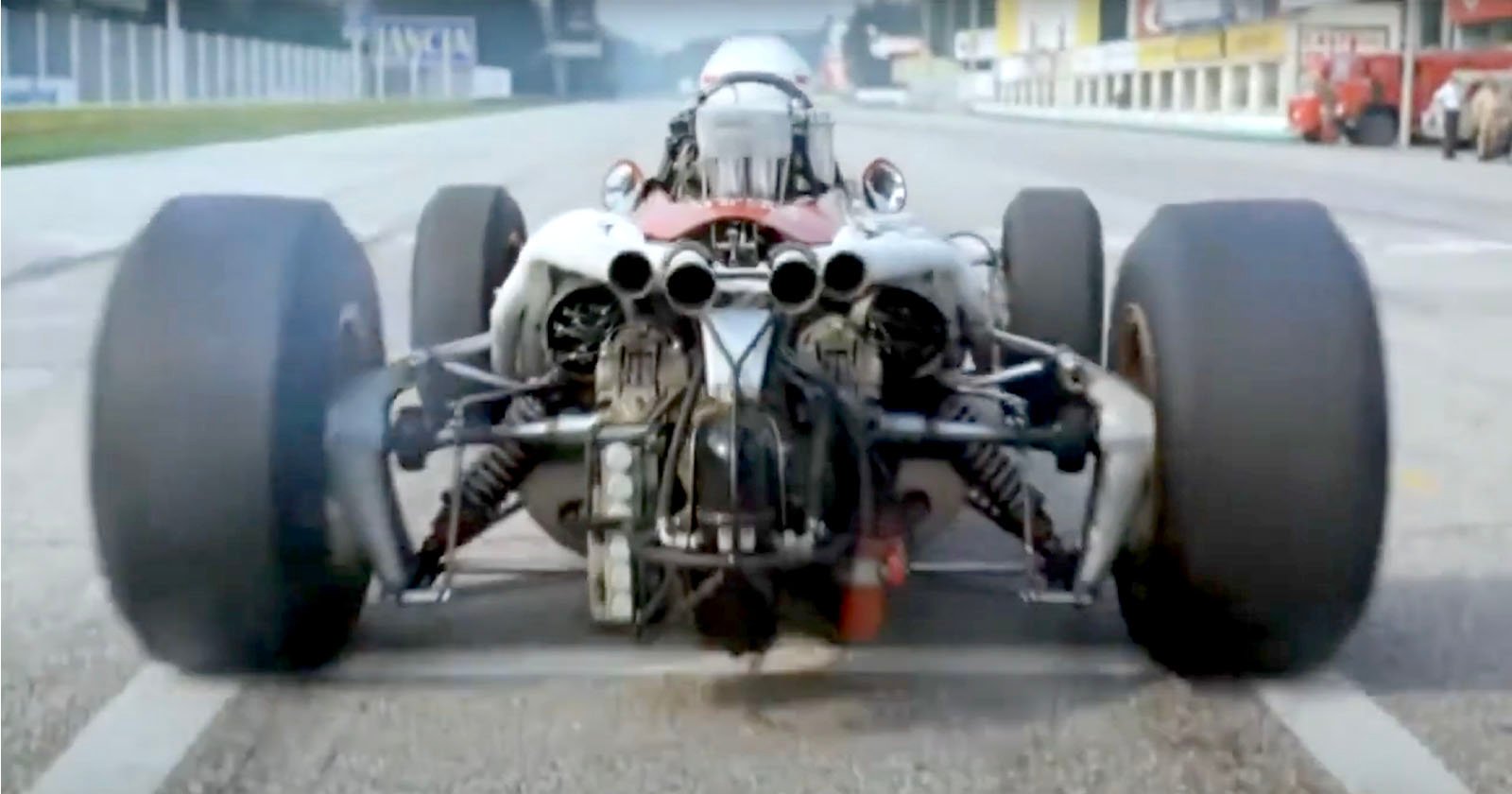 A rear view of a vintage Formula 1 race car on a track, showing wide tires, exposed suspension, and engine components, with some smoke rising from the left tire. A rear view of a vintage Formula 1 race car on a track, showing wide tires, exposed suspension, and engine components, with some smoke rising from the left tire.