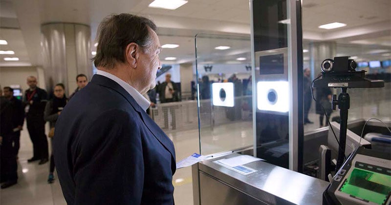 A man stands in front of a biometric scanner at an airport security checkpoint. Several people and staff are visible in the background of the brightly lit terminal. A man stands in front of a biometric scanner at an airport security checkpoint. Several people and staff are visible in the background of the brightly lit terminal.