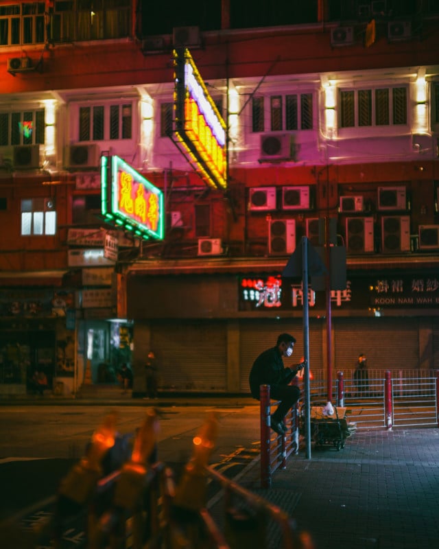 A nighttime street scene with a man sitting on a railing using his phone A nighttime street scene with a man sitting on a railing using his phone