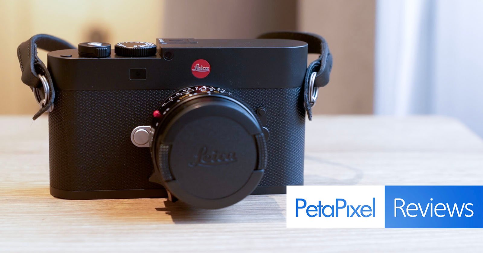 A black Leica camera with a lens cap is placed on a light wooden surface. The image features the PetaPixel Reviews logo in the bottom right corner. A black Leica camera with a lens cap is placed on a light wooden surface. The image features the PetaPixel Reviews logo in the bottom right corner.