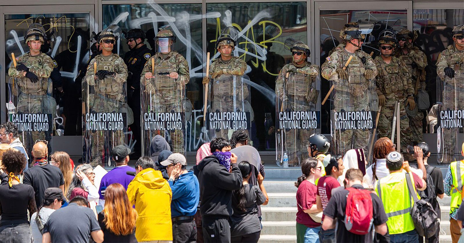 A line of California National Guard troops in riot gear stands in front of a building with glass doors covered in graffiti, facing a crowd of protesters gathered on the steps. A line of California National Guard troops in riot gear stands in front of a building with glass doors covered in graffiti, facing a crowd of protesters gathered on the steps.