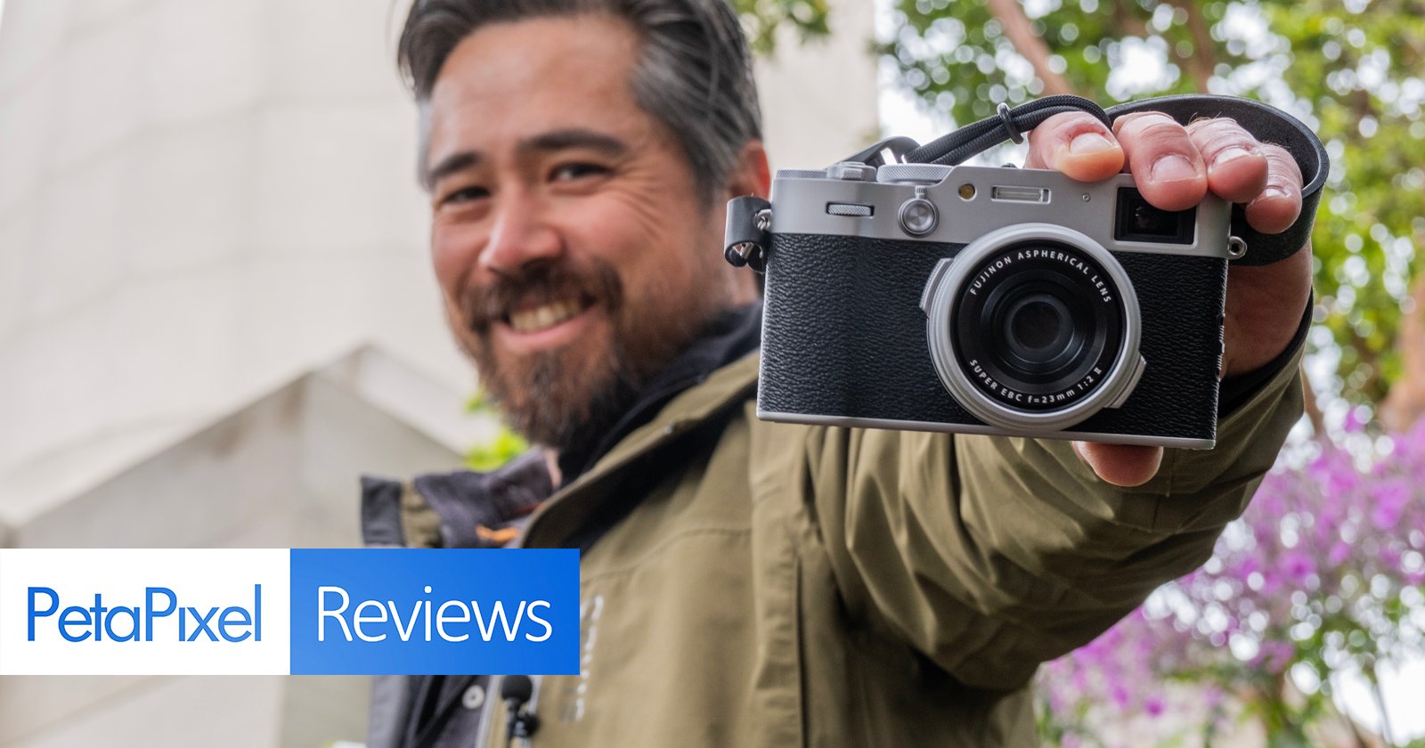 A smiling man holding a camera in front of him, focused on the lens, with petapixel reviews overlay text, in a garden setting.