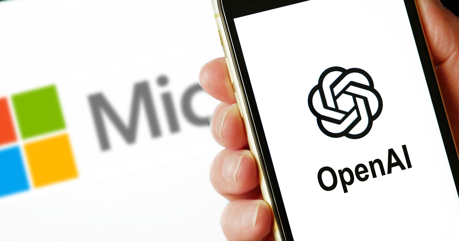 A person holds a smartphone displaying the openai logo, with a blurred microsoft logo visible in the background on another device. A person holds a smartphone displaying the openai logo, with a blurred microsoft logo visible in the background on another device.