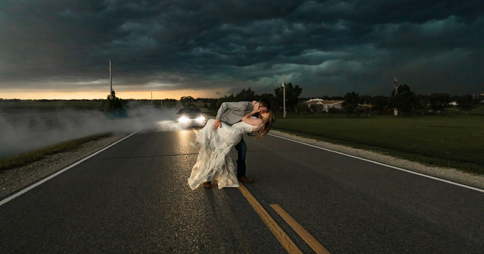 A couple shares a romantic kiss in the middle of a deserted road under a dark, stormy sky. The groom dips the bride as they embrace, while car headlights illuminate them from behind, creating a dramatic and cinematic atmosphere. A couple shares a romantic kiss in the middle of a deserted road under a dark, stormy sky. The groom dips the bride as they embrace, while car headlights illuminate them from behind, creating a dramatic and cinematic atmosphere.