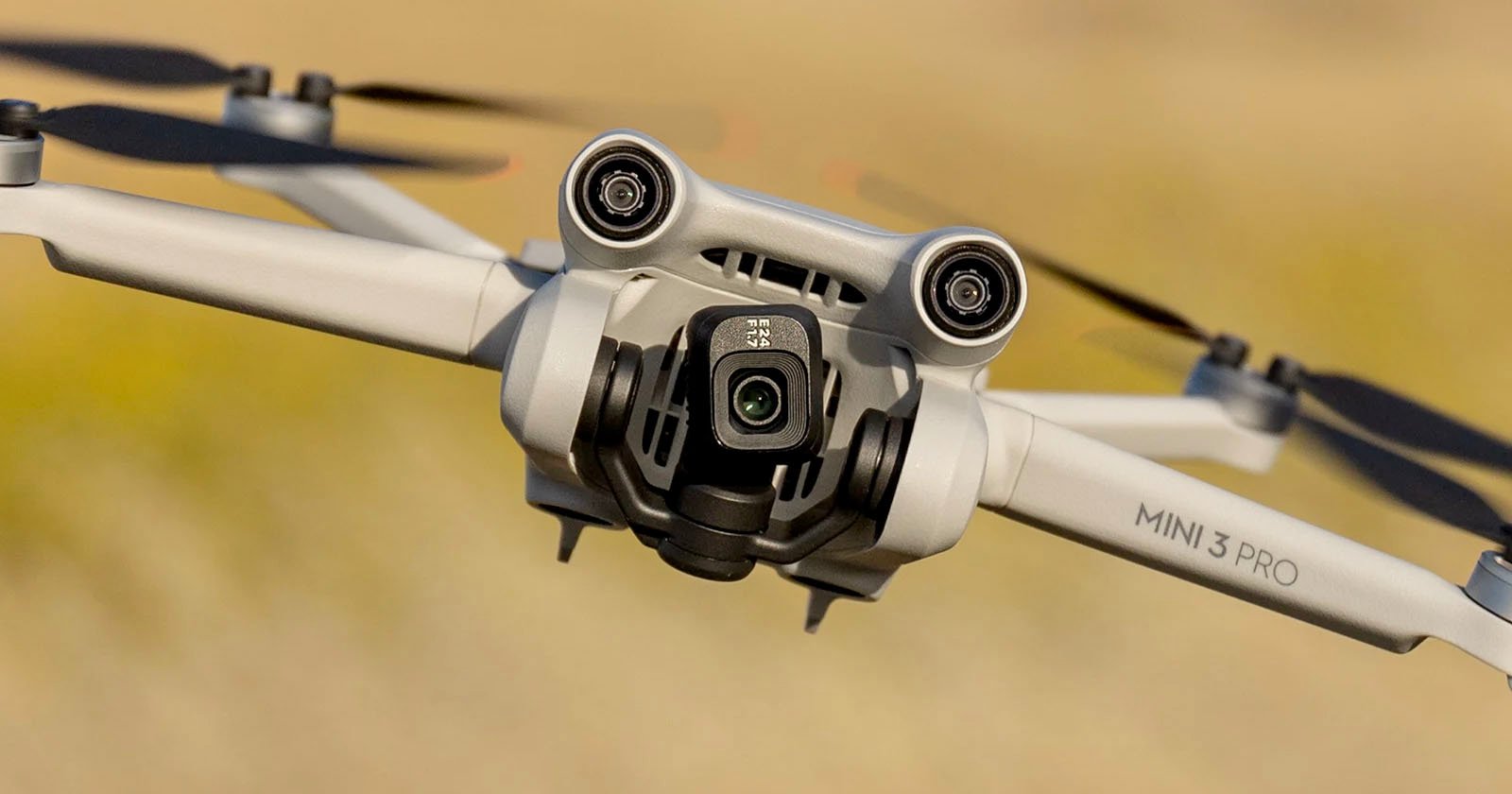 A close-up shot of the DJI Mini 3 Pro drone in flight, showing its front-facing camera, gimbal, and propellers. The background is a blurred, light brown landscape, highlighting the drone's details. The model name MINI 3 PRO is visible on one of its arms. A close-up shot of the DJI Mini 3 Pro drone in flight, showing its front-facing camera, gimbal, and propellers. The background is a blurred, light brown landscape, highlighting the drone's details. The model name MINI 3 PRO is visible on one of its arms.
