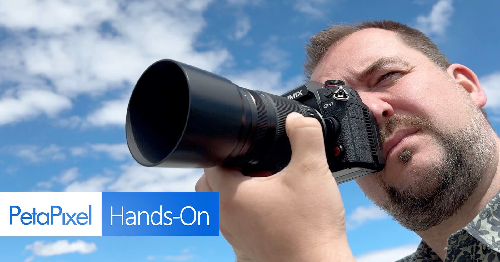 A man is looking through the viewfinder of a Panasonic GH7 camera pointed towards the sky. The background features a vivid blue sky with scattered clouds. The text PetaPixel Hands-On is displayed in the lower-left corner of the image. A man is looking through the viewfinder of a Panasonic GH7 camera pointed towards the sky. The background features a vivid blue sky with scattered clouds. The text PetaPixel Hands-On is displayed in the lower-left corner of the image.