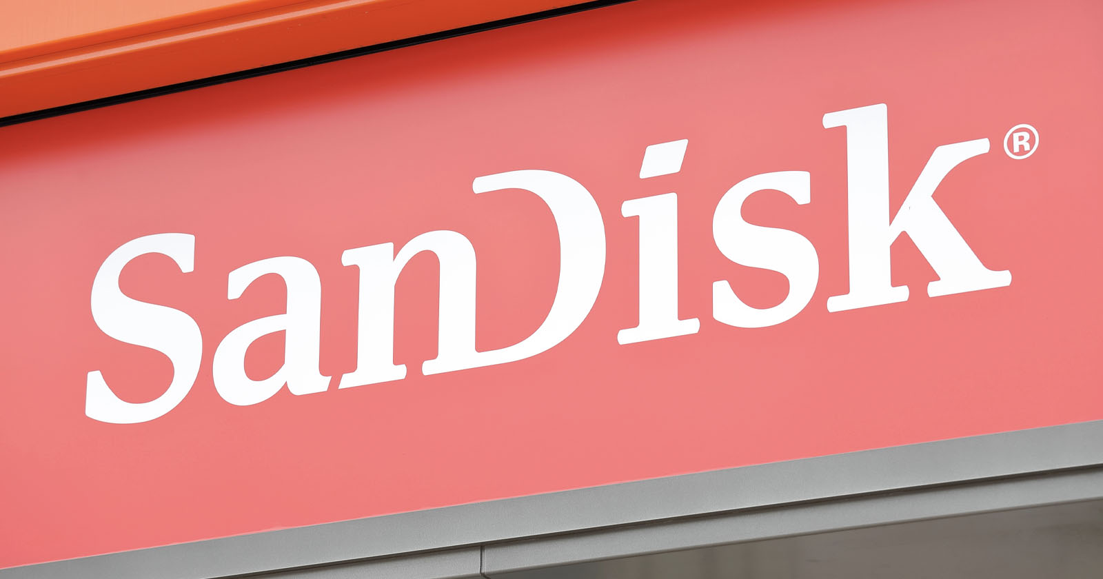 A red sign with the white text SanDisk is prominently displayed. The text is in a bold, modern font with a reserved trademark symbol at the top right corner of the letter k. A red sign with the white text SanDisk is prominently displayed. The text is in a bold, modern font with a reserved trademark symbol at the top right corner of the letter k.