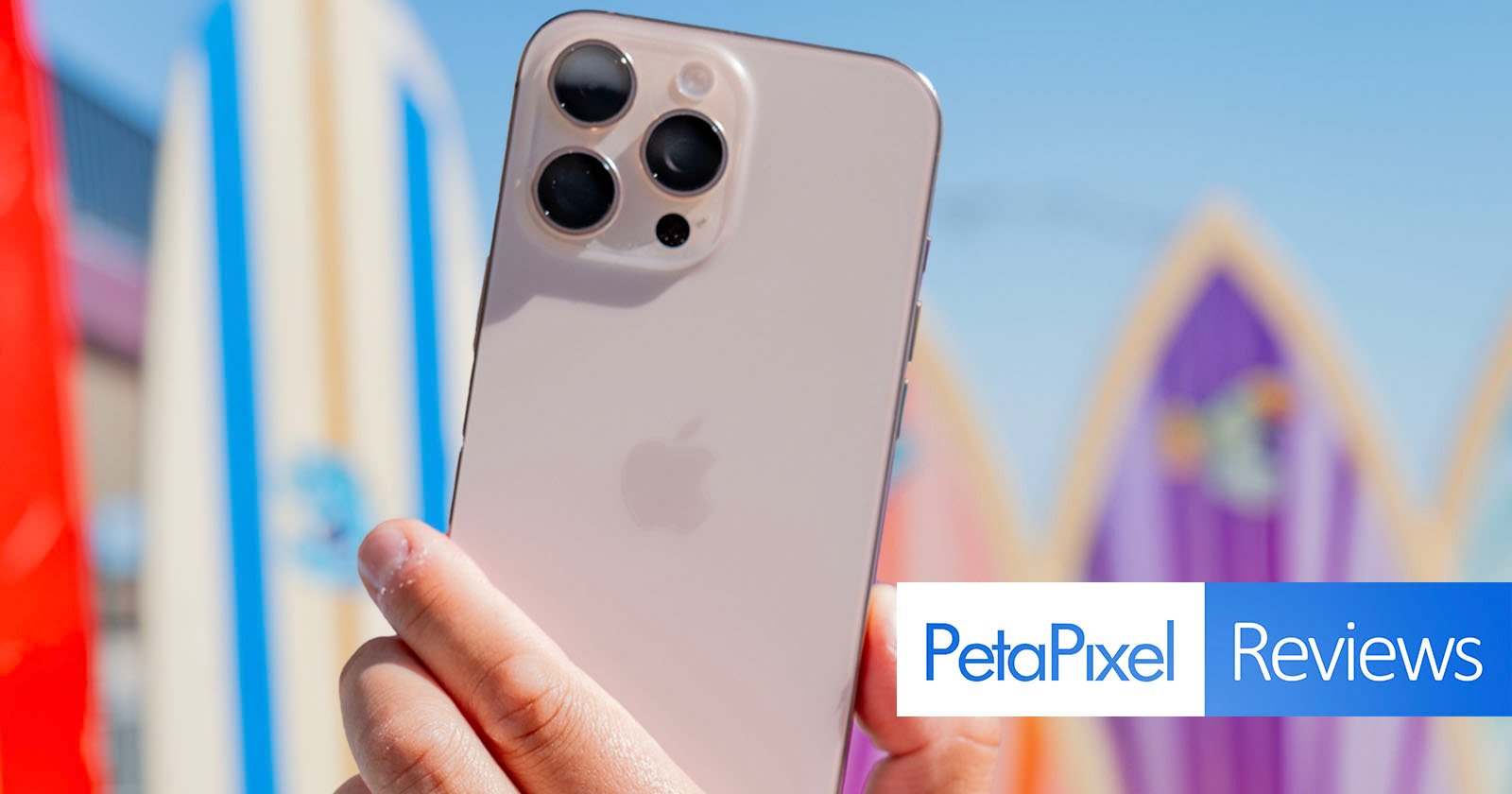 A person holds up an iPhone with a triple camera system in front of colorful surfboards. The back of the phone with the Apple logo is prominently visible. The image has a text overlay PetaPixel Reviews in the bottom right corner. A person holds up an iPhone with a triple camera system in front of colorful surfboards. The back of the phone with the Apple logo is prominently visible. The image has a text overlay PetaPixel Reviews in the bottom right corner.