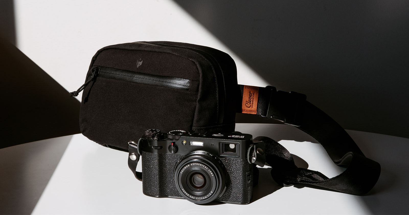 A black, vintage-style camera with a strap is placed on a white surface. Next to it is a black rectangular camera bag. Sunlight and shadows create a contrasting background. A black, vintage-style camera with a strap is placed on a white surface. Next to it is a black rectangular camera bag. Sunlight and shadows create a contrasting background.