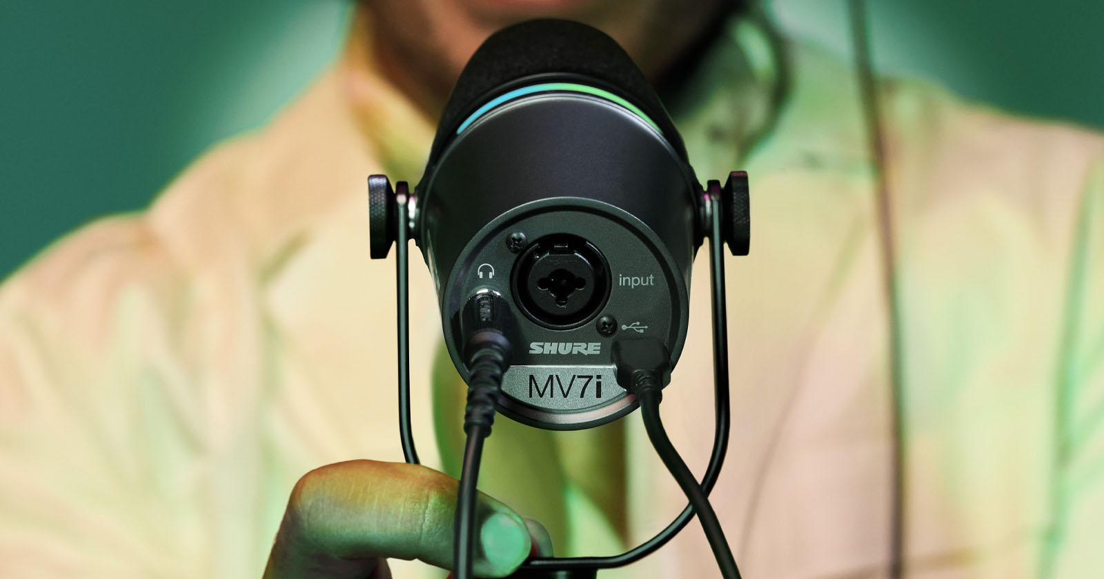 Close-up of a person holding a Shure MV7 microphone, showing the input connections at the back. The person is wearing a light-colored shirt, and the background is a soft green. Close-up of a person holding a Shure MV7 microphone, showing the input connections at the back. The person is wearing a light-colored shirt, and the background is a soft green.