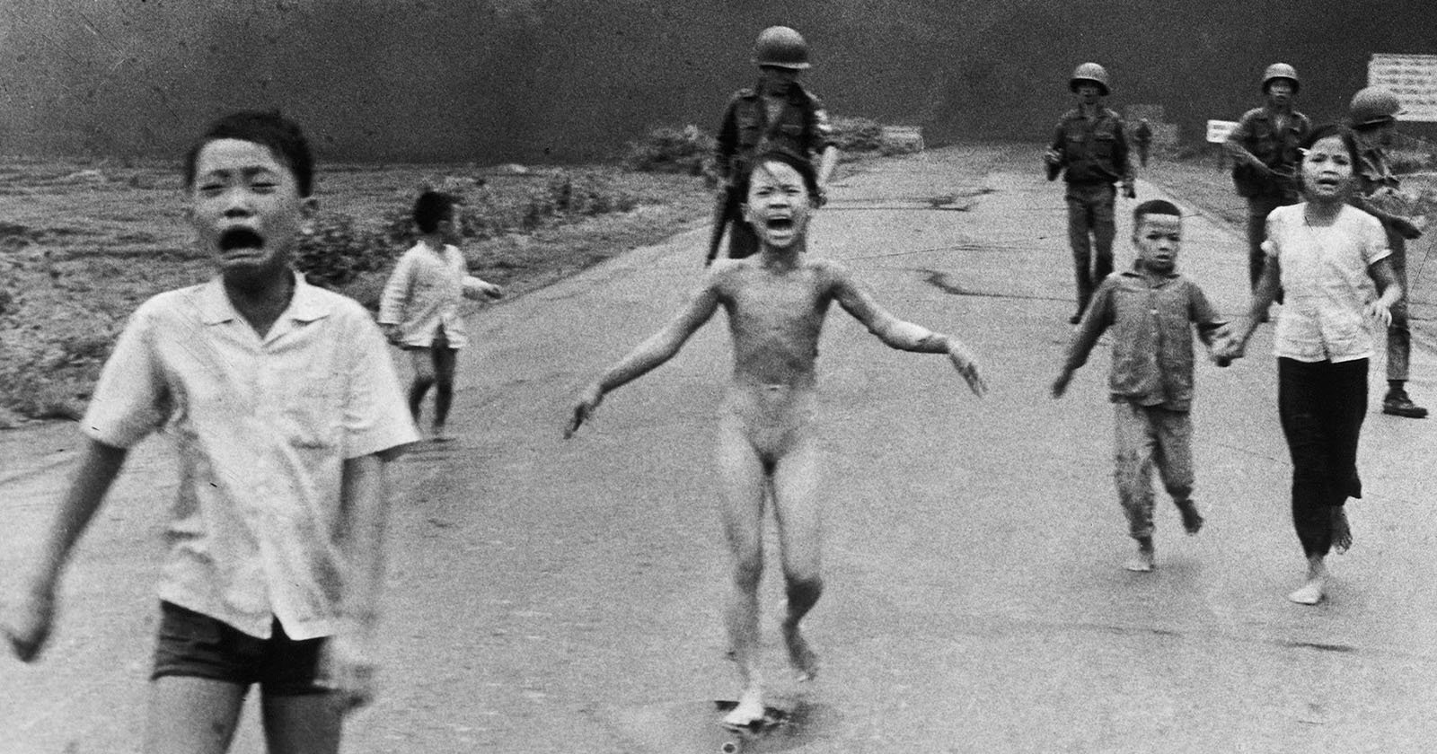 Children run down a road, appearing distressed, with soldiers walking behind them. A central child, without clothing, is crying. The background is a dark smoke-filled sky, conveying a sense of urgency and fear. Children run down a road, appearing distressed, with soldiers walking behind them. A central child, without clothing, is crying. The background is a dark smoke-filled sky, conveying a sense of urgency and fear.