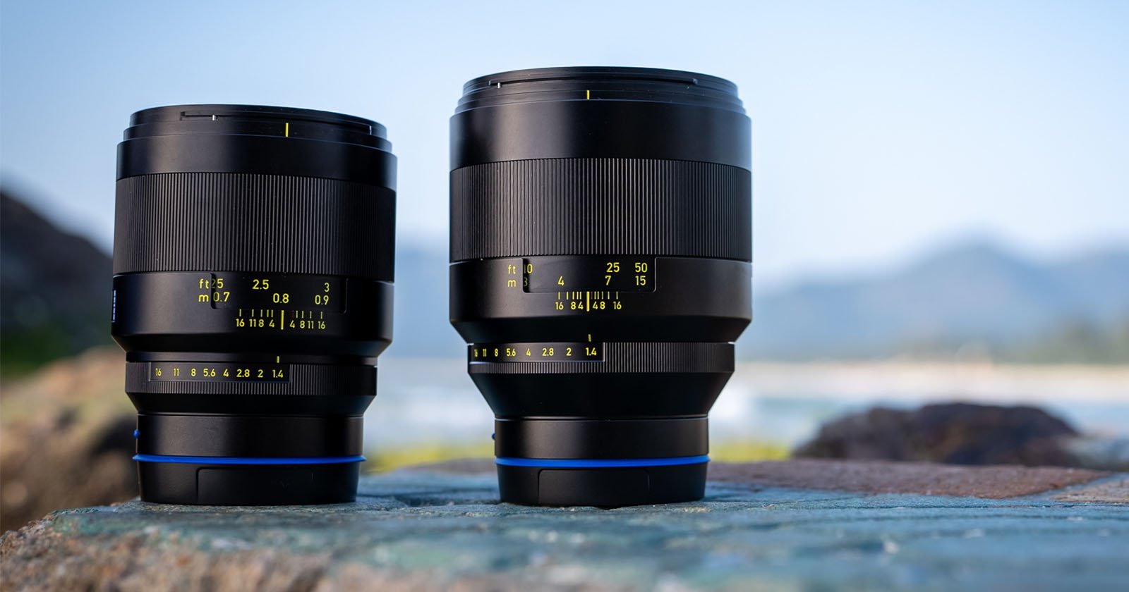 Two black camera lenses with yellow marking details stand on a stone surface. The background features a soft-focus landscape with mountains and a clear sky. Two black camera lenses with yellow marking details stand on a stone surface. The background features a soft-focus landscape with mountains and a clear sky.