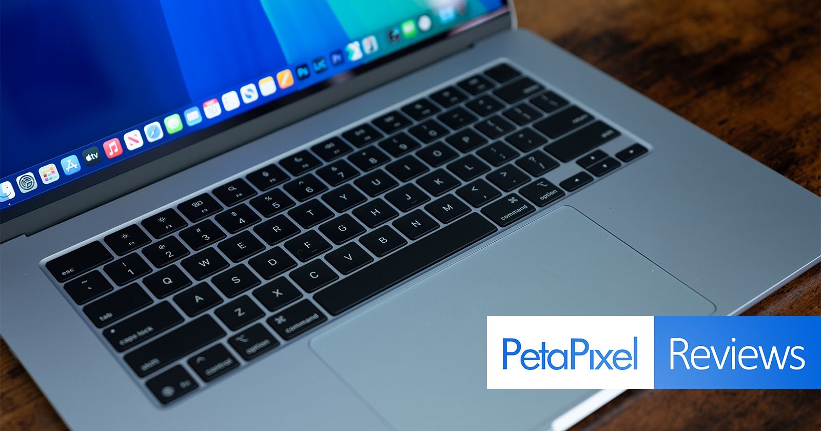 A silver laptop is open on a wooden surface, displaying a desktop screen with app icons. The keyboard and trackpad are visible. In the corner, there is an overlay with the words PetaPixel Reviews. A silver laptop is open on a wooden surface, displaying a desktop screen with app icons. The keyboard and trackpad are visible. In the corner, there is an overlay with the words PetaPixel Reviews.