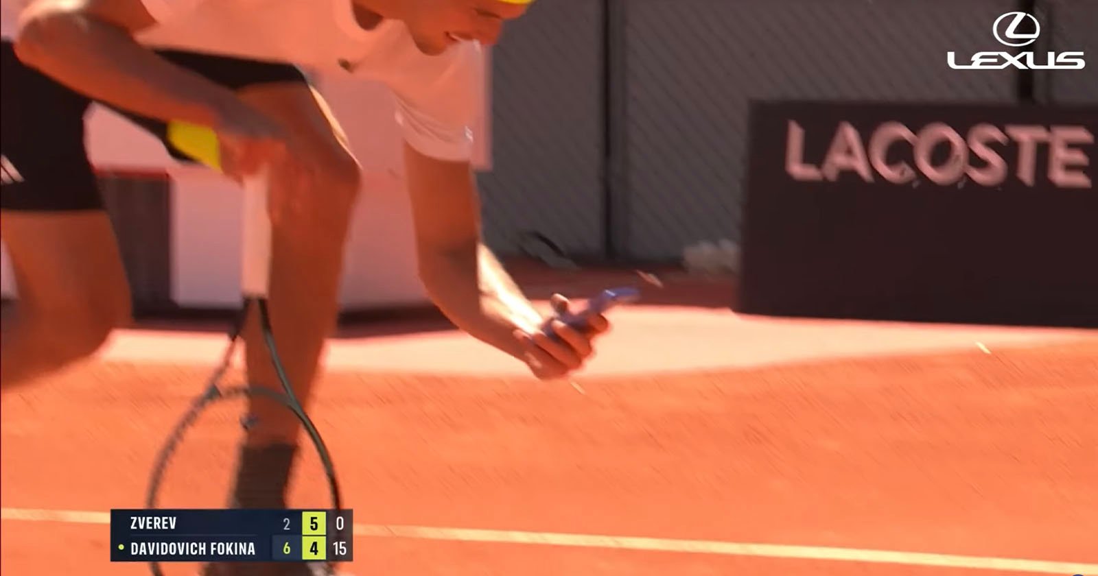 A tennis player in a white shirt and yellow headband bends down on a clay court, holding a phone and racket. Score graphics and the Lexus logo appear on the image. A tennis player in a white shirt and yellow headband bends down on a clay court, holding a phone and racket. Score graphics and the Lexus logo appear on the image.