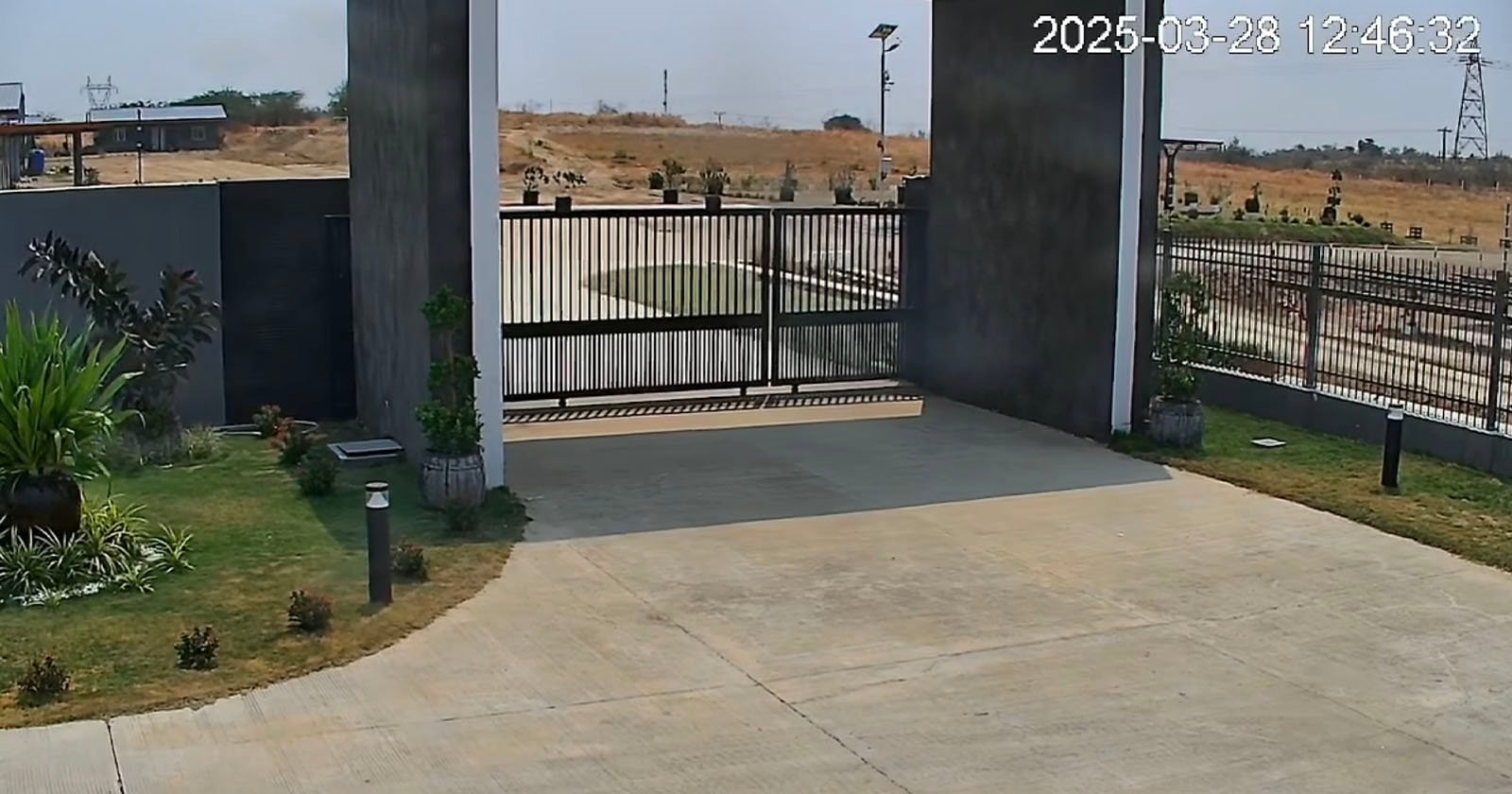 A closed black metal gate blocks the entrance to a paved driveway, surrounded by landscaped grass and plants. The background shows a dry, open field under a clear sky. The date and time read 2025-03-28 12:46:32. A closed black metal gate blocks the entrance to a paved driveway, surrounded by landscaped grass and plants. The background shows a dry, open field under a clear sky. The date and time read 2025-03-28 12:46:32.