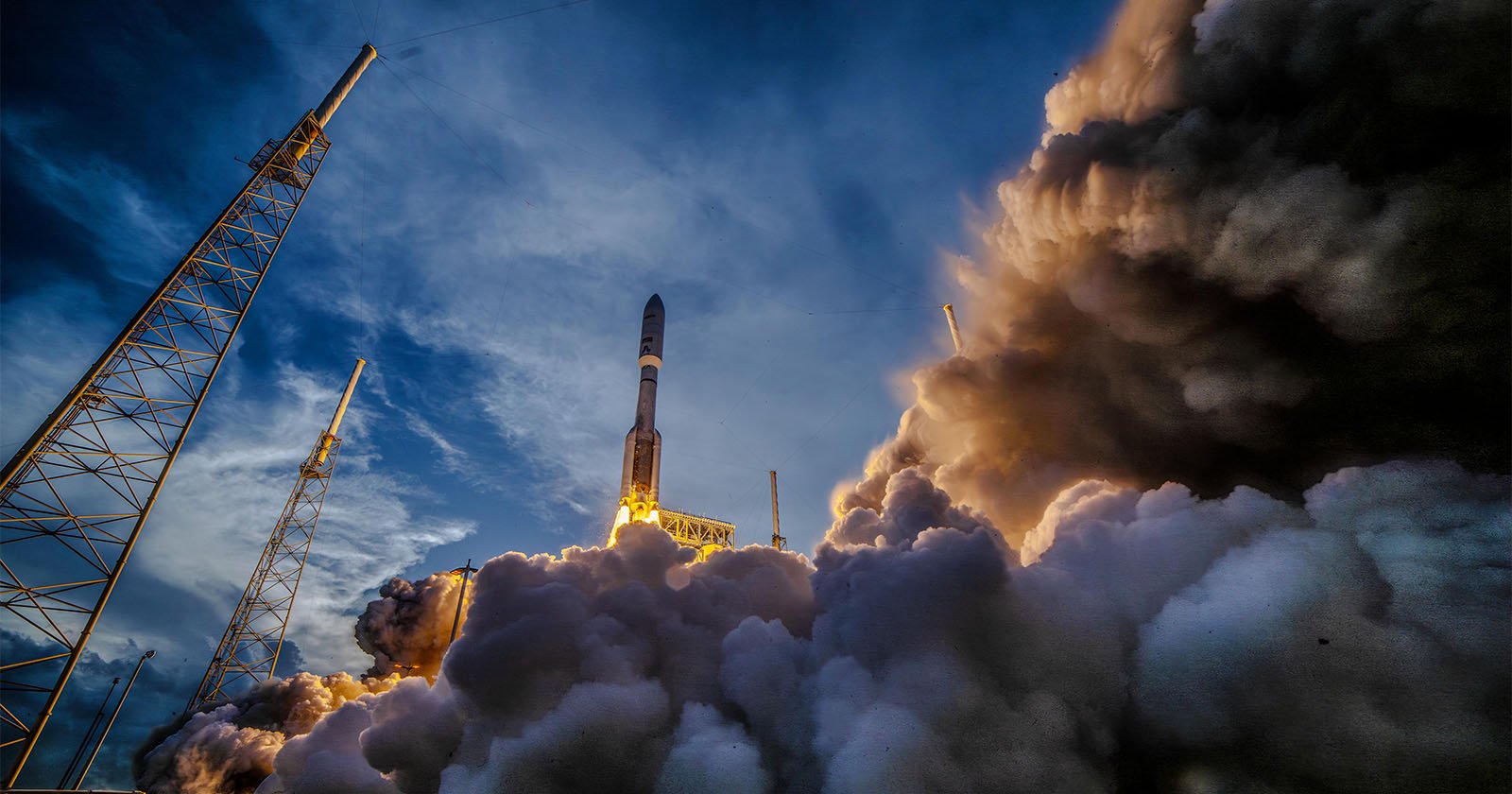 A rocket launches into the sky, surrounded by billowing clouds of smoke and steam, with a dramatic blue and cloudy evening sky in the background and launch towers on either side. A rocket launches into the sky, surrounded by billowing clouds of smoke and steam, with a dramatic blue and cloudy evening sky in the background and launch towers on either side.