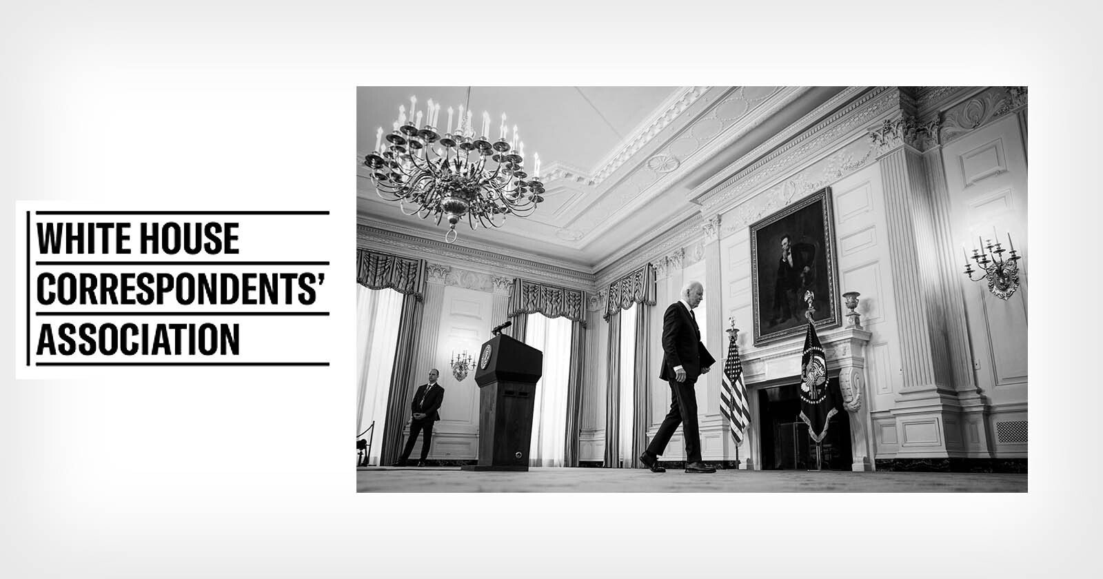 Black and white photo of a man walking past a fireplace and portrait in an ornate room with chandeliers; podium on left. Text on left reads White House Correspondents' Association. Black and white photo of a man walking past a fireplace and portrait in an ornate room with chandeliers; podium on left. Text on left reads White House Correspondents' Association.