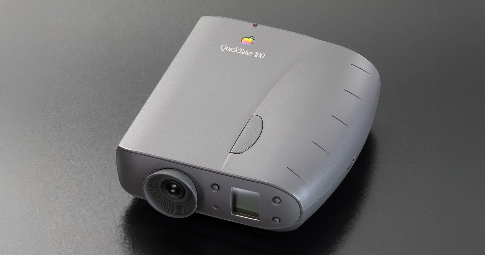 A gray Apple QuickTake 100 digital camera with a rounded body, lens on the front, and Apple logo on top, placed on a sleek dark surface. A gray Apple QuickTake 100 digital camera with a rounded body, lens on the front, and Apple logo on top, placed on a sleek dark surface.