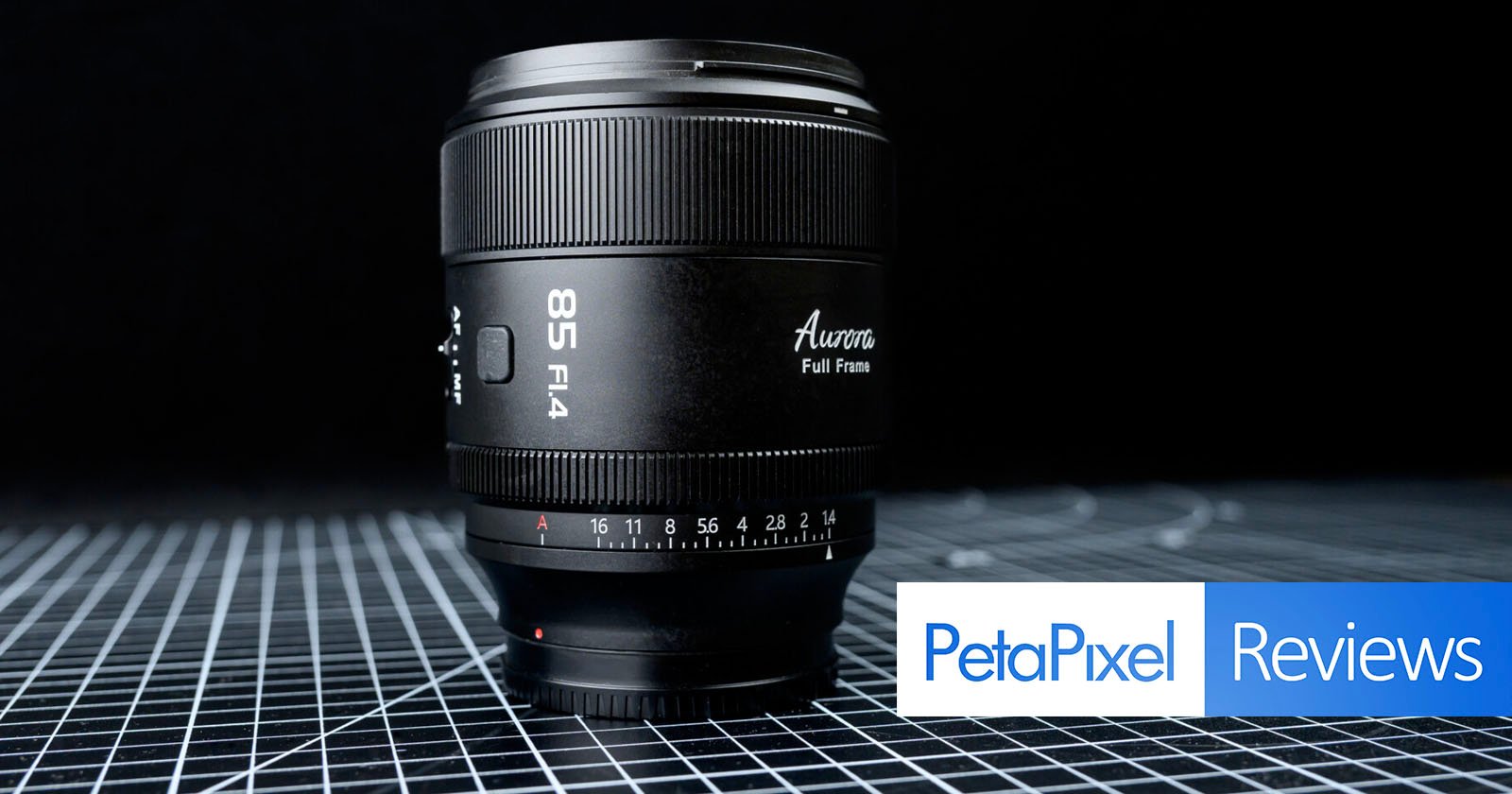 A black 85mm f/1.4 camera lens sits upright on a grid-patterned surface, with the text Petapixel Reviews in blue and white in the lower right corner. A black 85mm f/1.4 camera lens sits upright on a grid-patterned surface, with the text Petapixel Reviews in blue and white in the lower right corner.
