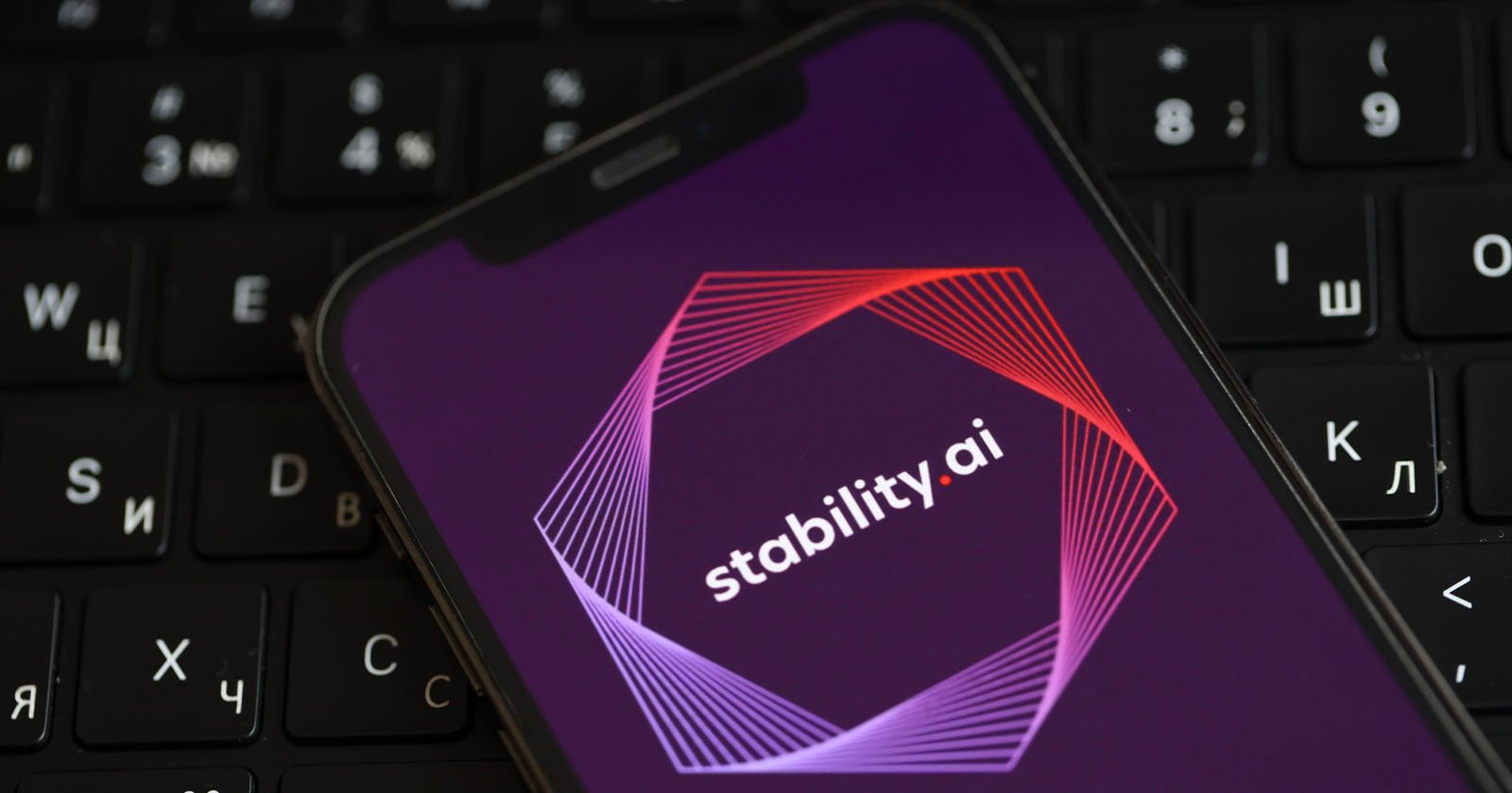 A smartphone displays the Stability.ai logo on its screen, featuring geometric lines in pink and red around the name. The phone rests on a black keyboard with Cyrillic and Latin characters. A smartphone displays the Stability.ai logo on its screen, featuring geometric lines in pink and red around the name. The phone rests on a black keyboard with Cyrillic and Latin characters.
