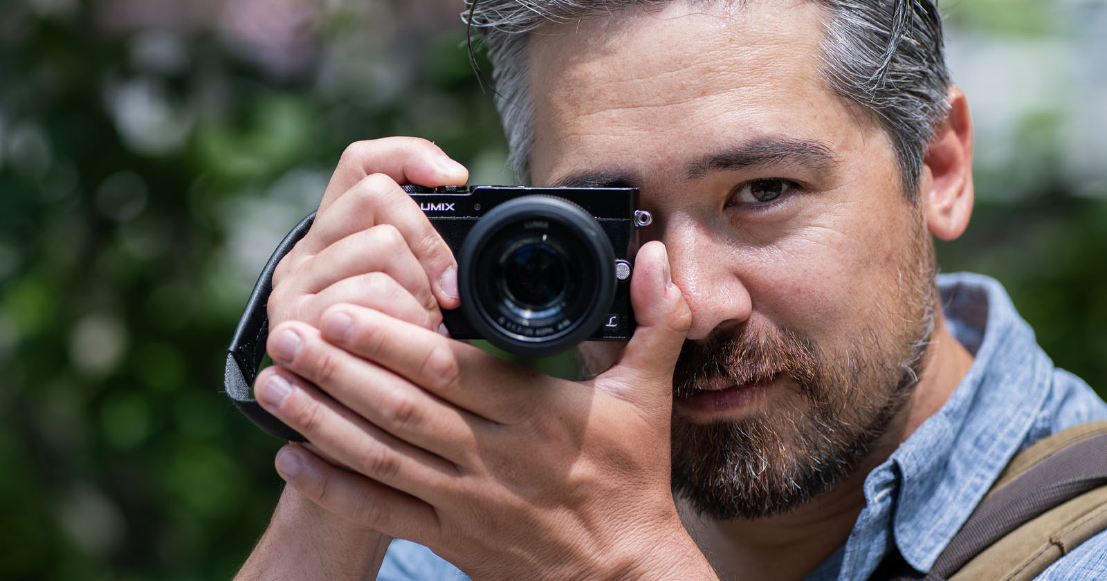 A man with gray hair and a beard is holding a camera up to his face, preparing to take a photo. He is outdoors, wearing a blue shirt, and the background is blurred greenery. A man with gray hair and a beard is holding a camera up to his face, preparing to take a photo. He is outdoors, wearing a blue shirt, and the background is blurred greenery.