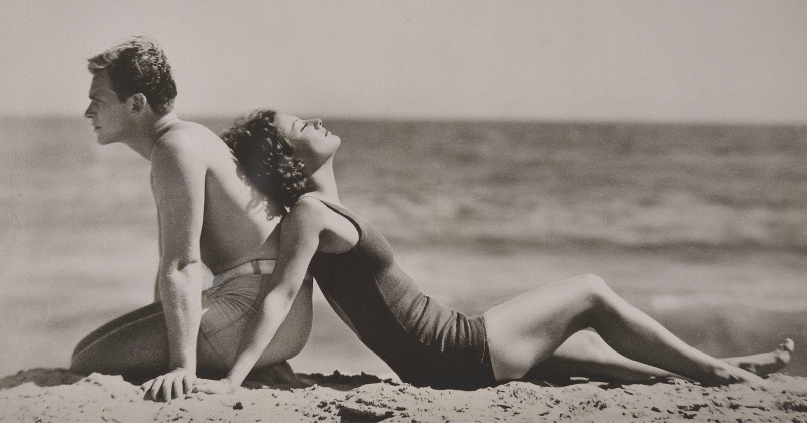 A man and woman in vintage swimsuits sit back to back on a sandy beach, facing the ocean. The woman leans back with her face tilted upward, eyes closed, enjoying the sun. The photo is in black and white. A man and woman in vintage swimsuits sit back to back on a sandy beach, facing the ocean. The woman leans back with her face tilted upward, eyes closed, enjoying the sun. The photo is in black and white.