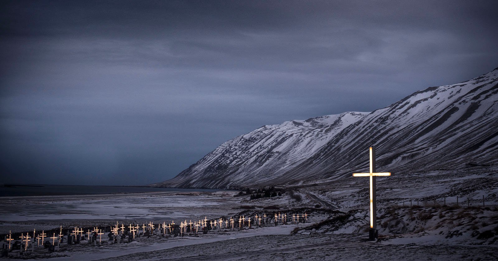 A snowy, coastal graveyard with rows of illuminated crosses at dusk, set against dramatic mountains and a cloudy sky. One large glowing cross stands prominently in the foreground. A snowy, coastal graveyard with rows of illuminated crosses at dusk, set against dramatic mountains and a cloudy sky. One large glowing cross stands prominently in the foreground.