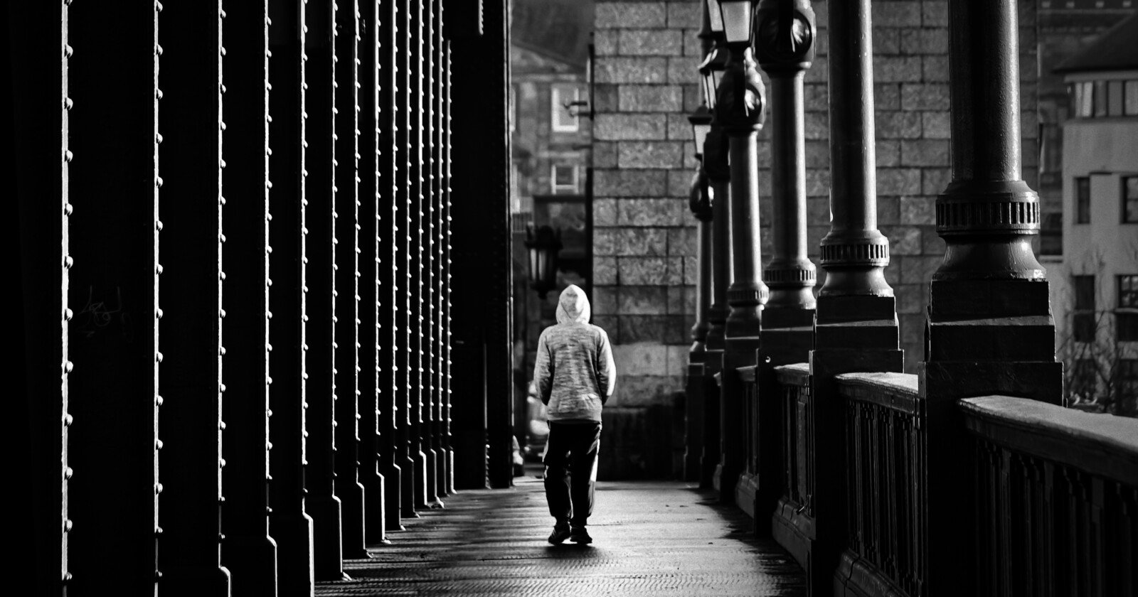 A person wearing a hooded jacket walks alone on a bridge, framed by tall columns and lamps, in a black-and-white urban setting. A person wearing a hooded jacket walks alone on a bridge, framed by tall columns and lamps, in a black-and-white urban setting.