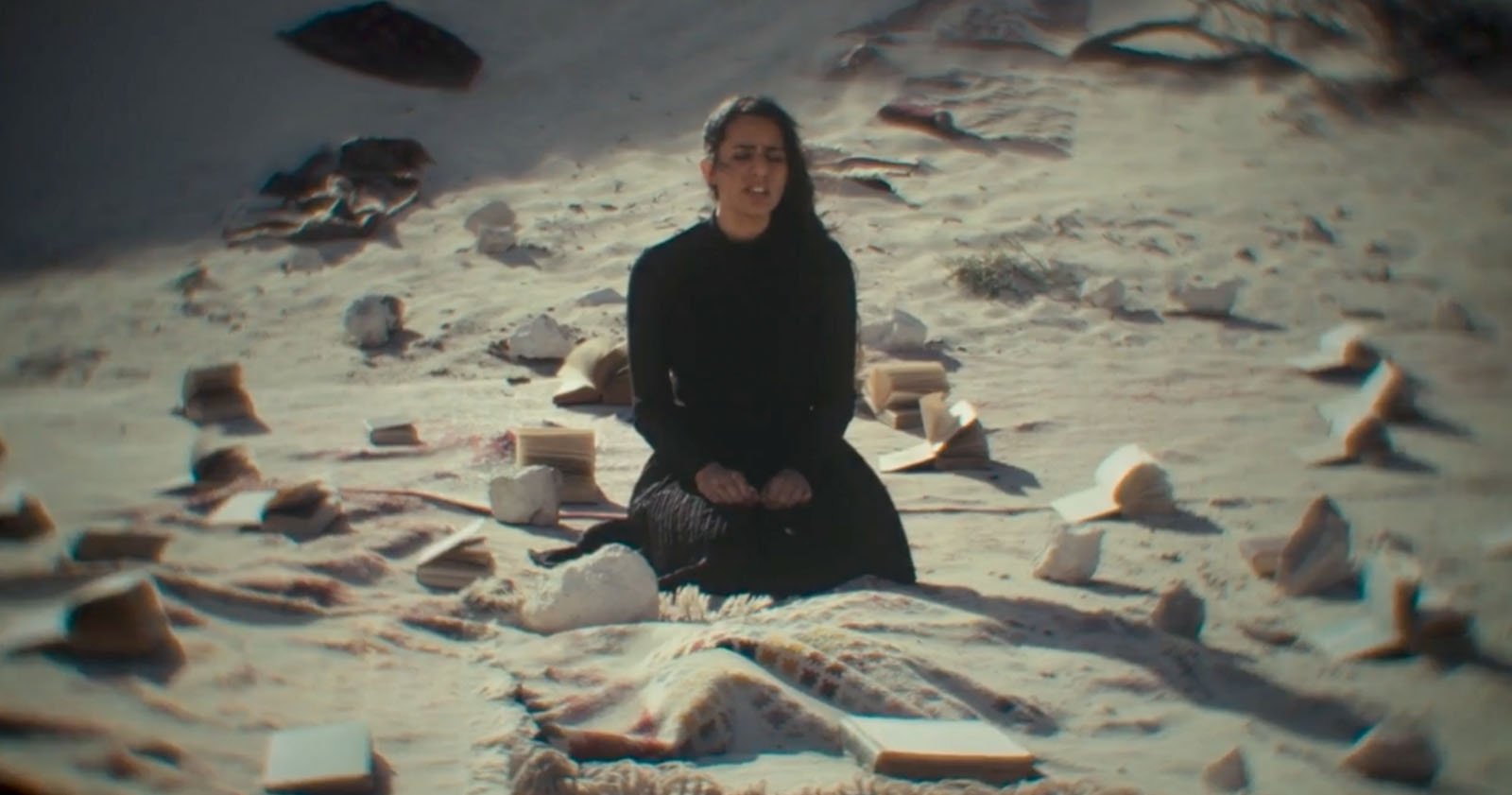 A woman in black sits cross-legged on a blanket in the sand, surrounded by open books and large stones scattered around her under bright daylight. A woman in black sits cross-legged on a blanket in the sand, surrounded by open books and large stones scattered around her under bright daylight.