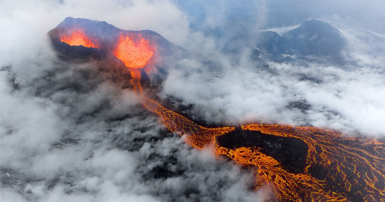 Aerial view of an erupting volcano with bright orange lava flowing down its slopes, surrounded by thick clouds and dark volcanic terrain. Aerial view of an erupting volcano with bright orange lava flowing down its slopes, surrounded by thick clouds and dark volcanic terrain.