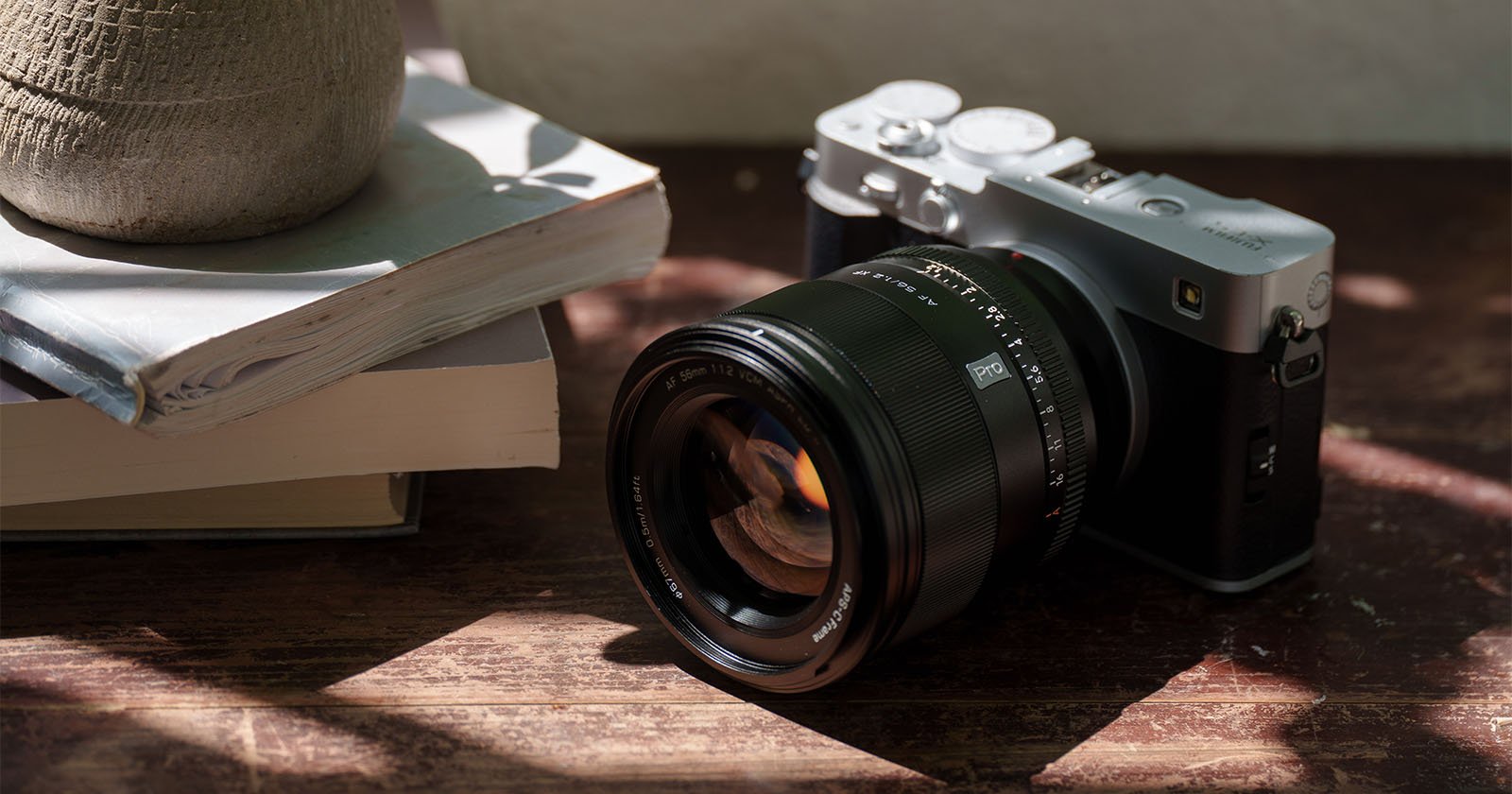 A digital camera with a large lens rests on a wooden surface next to a stack of books and a textured pot, with dappled sunlight casting shadows across the scene. A digital camera with a large lens rests on a wooden surface next to a stack of books and a textured pot, with dappled sunlight casting shadows across the scene.