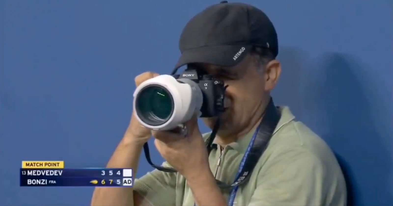 A photographer wearing a cap and green shirt takes a picture with a large camera at a tennis match. The scoreboard shows Medvedev vs. Bonzi, with Bonzi at match point. A photographer wearing a cap and green shirt takes a picture with a large camera at a tennis match. The scoreboard shows Medvedev vs. Bonzi, with Bonzi at match point.