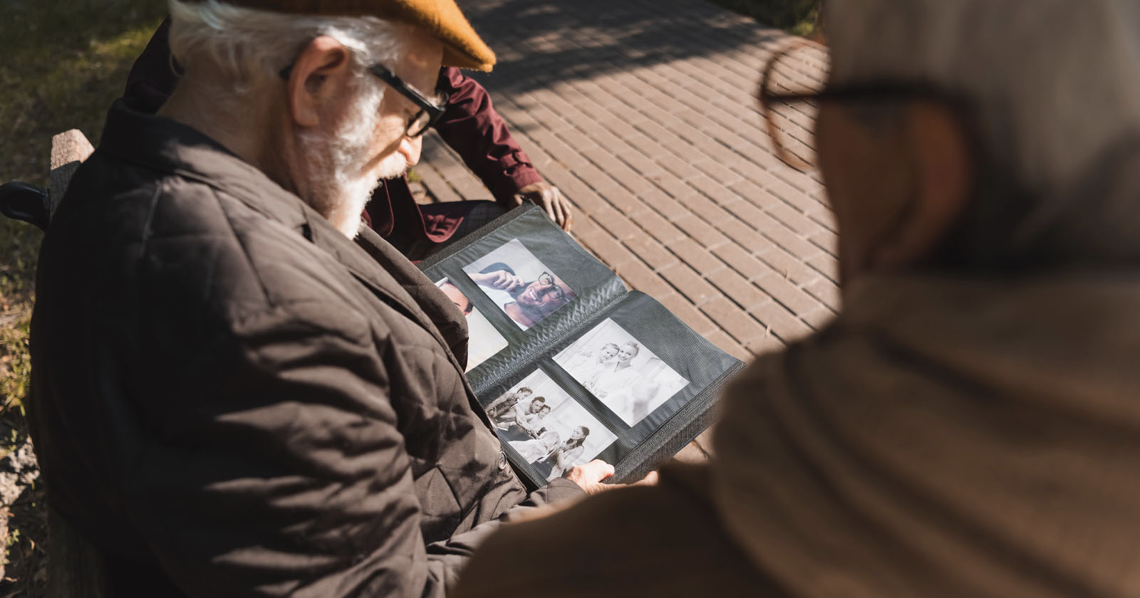 Two elderly people sit on a bench outdoors, looking at a photo album filled with black-and-white pictures. Sunlight casts shadows on the wooden path beside them. Two elderly people sit on a bench outdoors, looking at a photo album filled with black-and-white pictures. Sunlight casts shadows on the wooden path beside them.