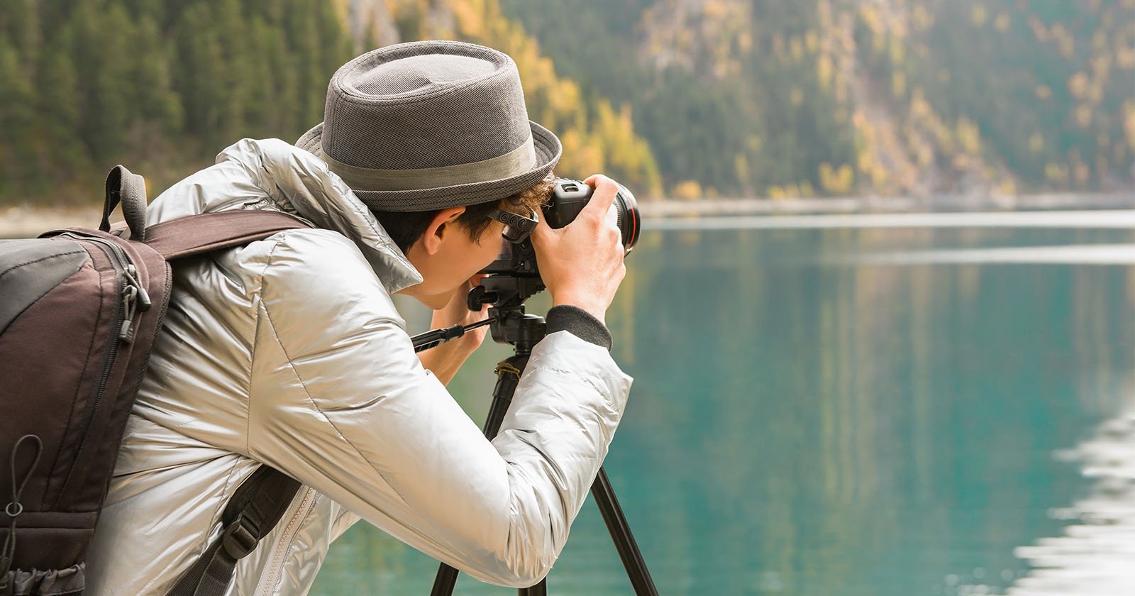 A person wearing a gray hat and jacket takes a photo with a camera on a tripod beside a calm lake, with trees and mountains in the background. A person wearing a gray hat and jacket takes a photo with a camera on a tripod beside a calm lake, with trees and mountains in the background.