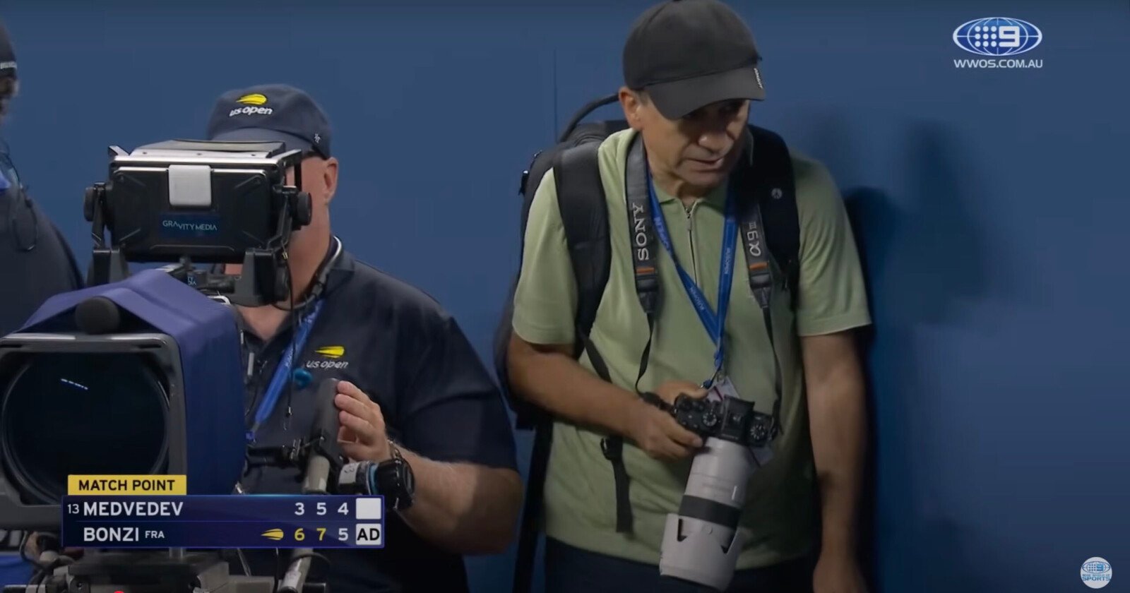 Two photographers stand courtside at a tennis match. One operates a large TV camera, while the other holds a professional camera with a telephoto lens. A scoreboard shows Medvedev vs. Bonzi with Bonzi at match point. Two photographers stand courtside at a tennis match. One operates a large TV camera, while the other holds a professional camera with a telephoto lens. A scoreboard shows Medvedev vs. Bonzi with Bonzi at match point.