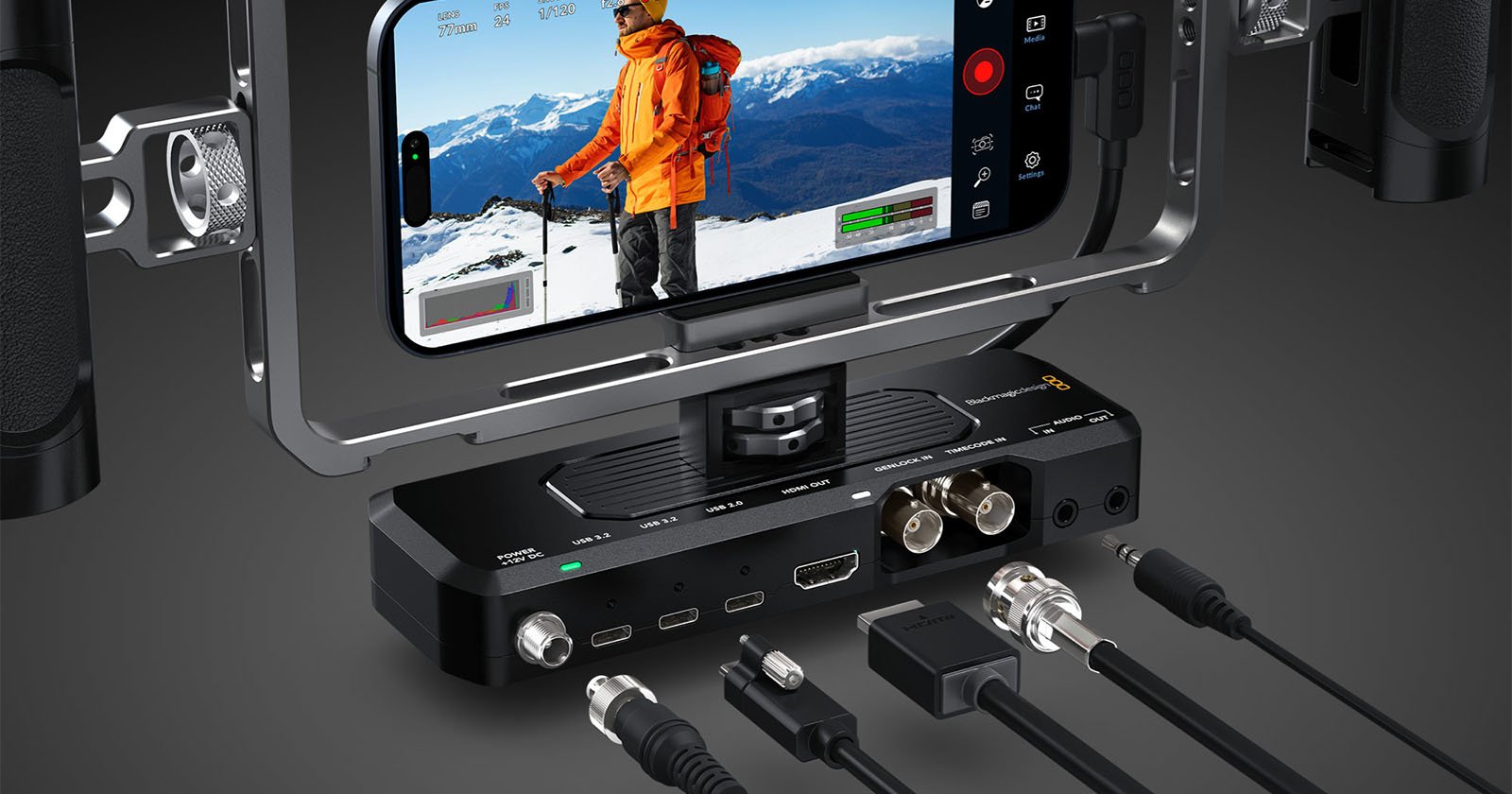 A smartphone mounted in a filming rig displays a person in an orange jacket on a snowy mountain. Below, various video and audio cables are connected to a compact black device with multiple ports and controls. A smartphone mounted in a filming rig displays a person in an orange jacket on a snowy mountain. Below, various video and audio cables are connected to a compact black device with multiple ports and controls.