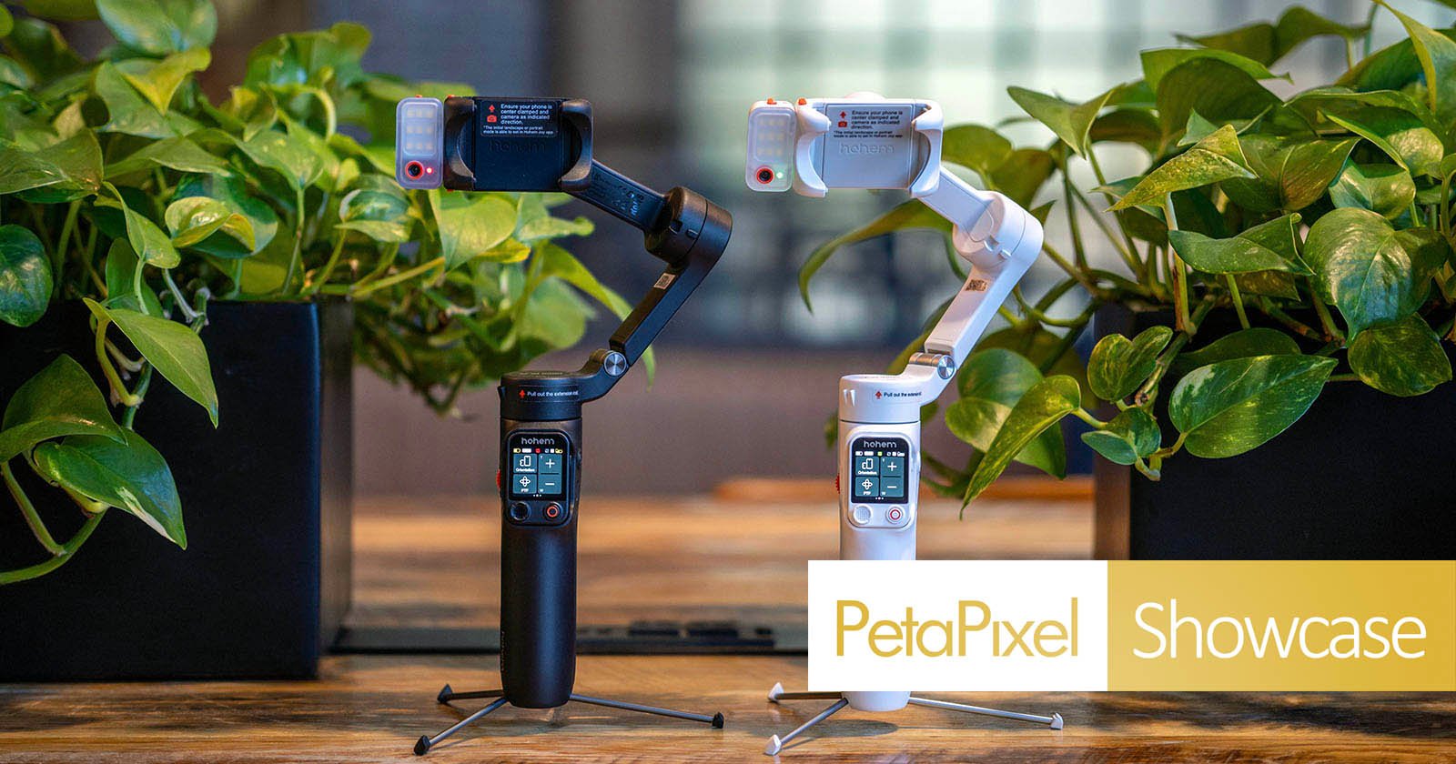 Two handheld gimbals, one black and one white, are standing on a wooden table surrounded by green plants. Both have small screens and are branded Hohem. PetaPixel Showcase text is displayed in the corner. Two handheld gimbals, one black and one white, are standing on a wooden table surrounded by green plants. Both have small screens and are branded Hohem. PetaPixel Showcase text is displayed in the corner.