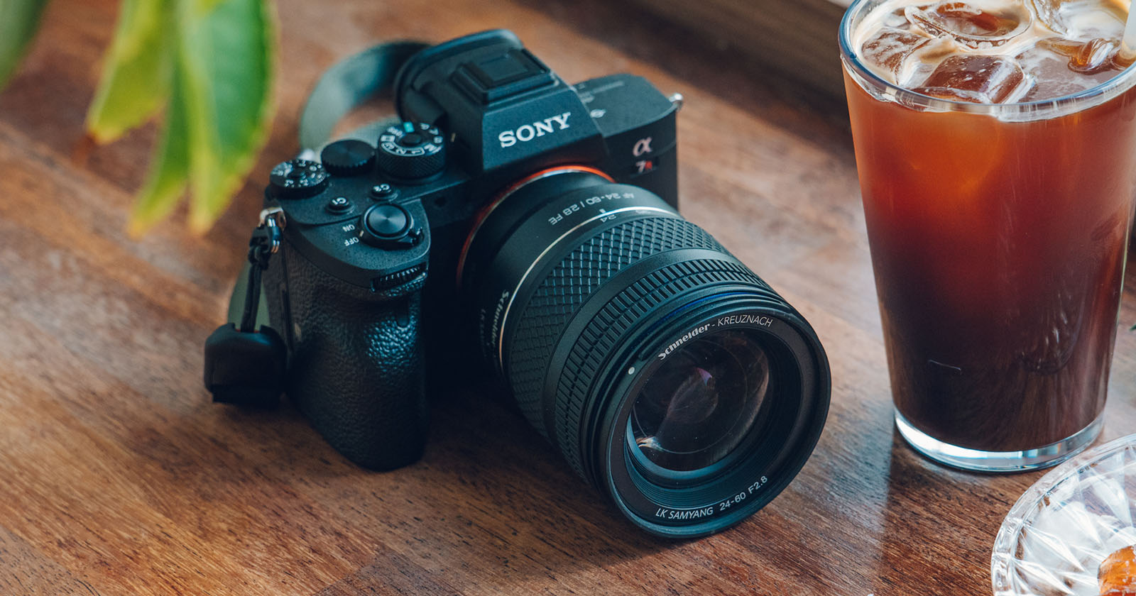 A Sony camera with a large lens rests on a wooden table next to a glass of iced coffee. Green leaves are visible in the background, and the scene has natural lighting. A Sony camera with a large lens rests on a wooden table next to a glass of iced coffee. Green leaves are visible in the background, and the scene has natural lighting.
