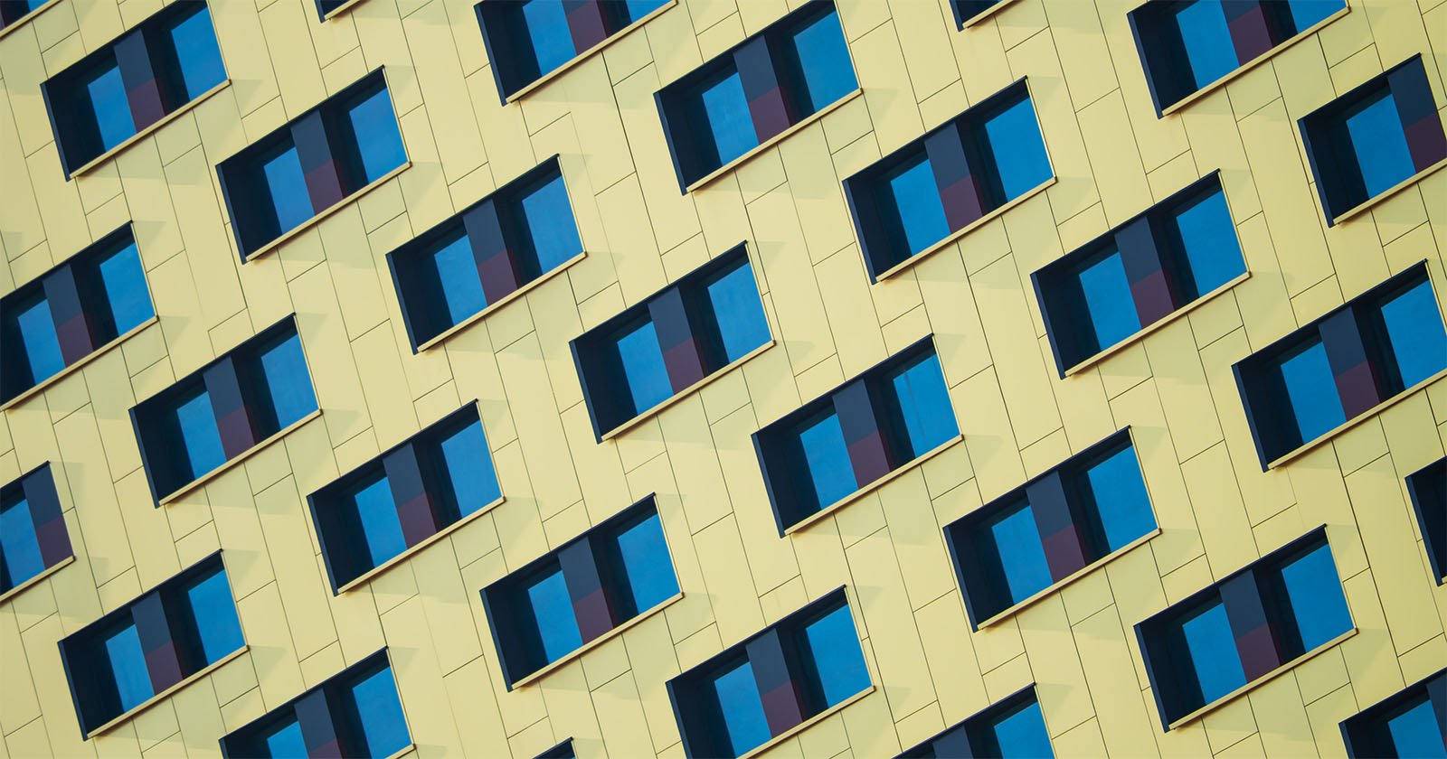 A close-up view of a modern building facade with a diagonal pattern of yellow panels and blue window shades, creating a geometric, repetitive visual effect. A close-up view of a modern building facade with a diagonal pattern of yellow panels and blue window shades, creating a geometric, repetitive visual effect.