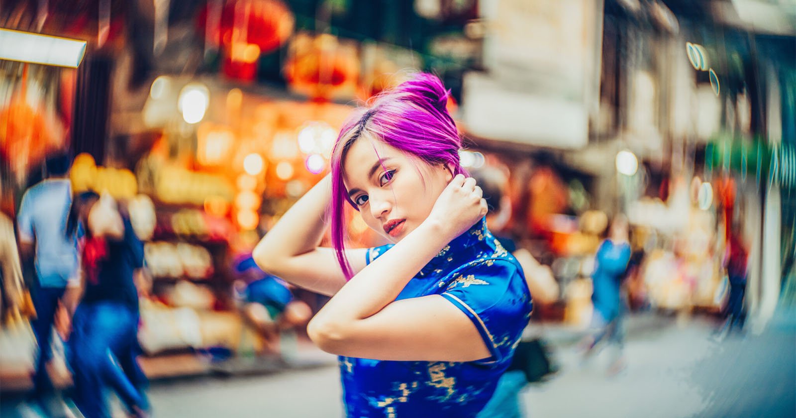 A young woman with vibrant purple hair, wearing a blue patterned dress, poses with her hands near her head on a bustling, colorful city street with blurred people and lights in the background. A young woman with vibrant purple hair, wearing a blue patterned dress, poses with her hands near her head on a bustling, colorful city street with blurred people and lights in the background.