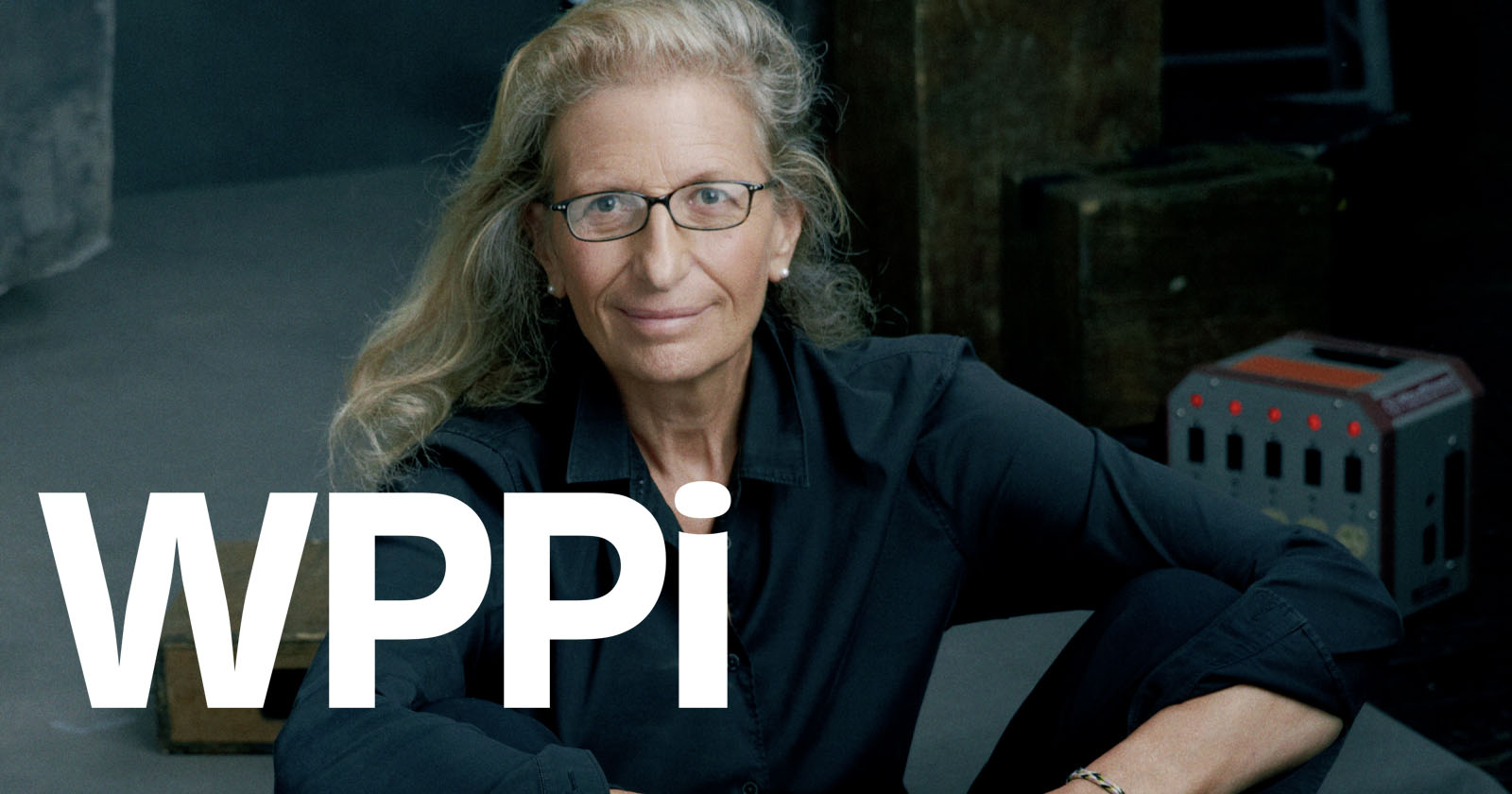 A woman with long, wavy blonde hair and glasses, wearing a black shirt, sits with her arms resting on her knees. Large white letters WPPI are overlaid on the image. A woman with long, wavy blonde hair and glasses, wearing a black shirt, sits with her arms resting on her knees. Large white letters WPPI are overlaid on the image.