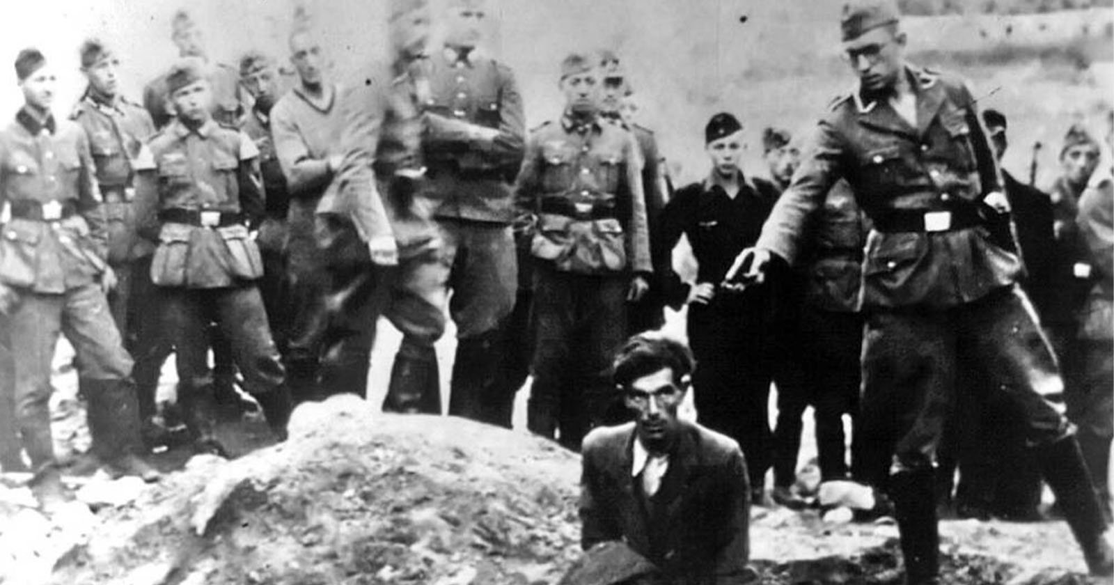 A group of uniformed soldiers stand outdoors as one soldier points a gun at a kneeling man with glasses, suggesting an execution scene; other men observe somberly in the background. The image is in black and white. A group of uniformed soldiers stand outdoors as one soldier points a gun at a kneeling man with glasses, suggesting an execution scene; other men observe somberly in the background. The image is in black and white.