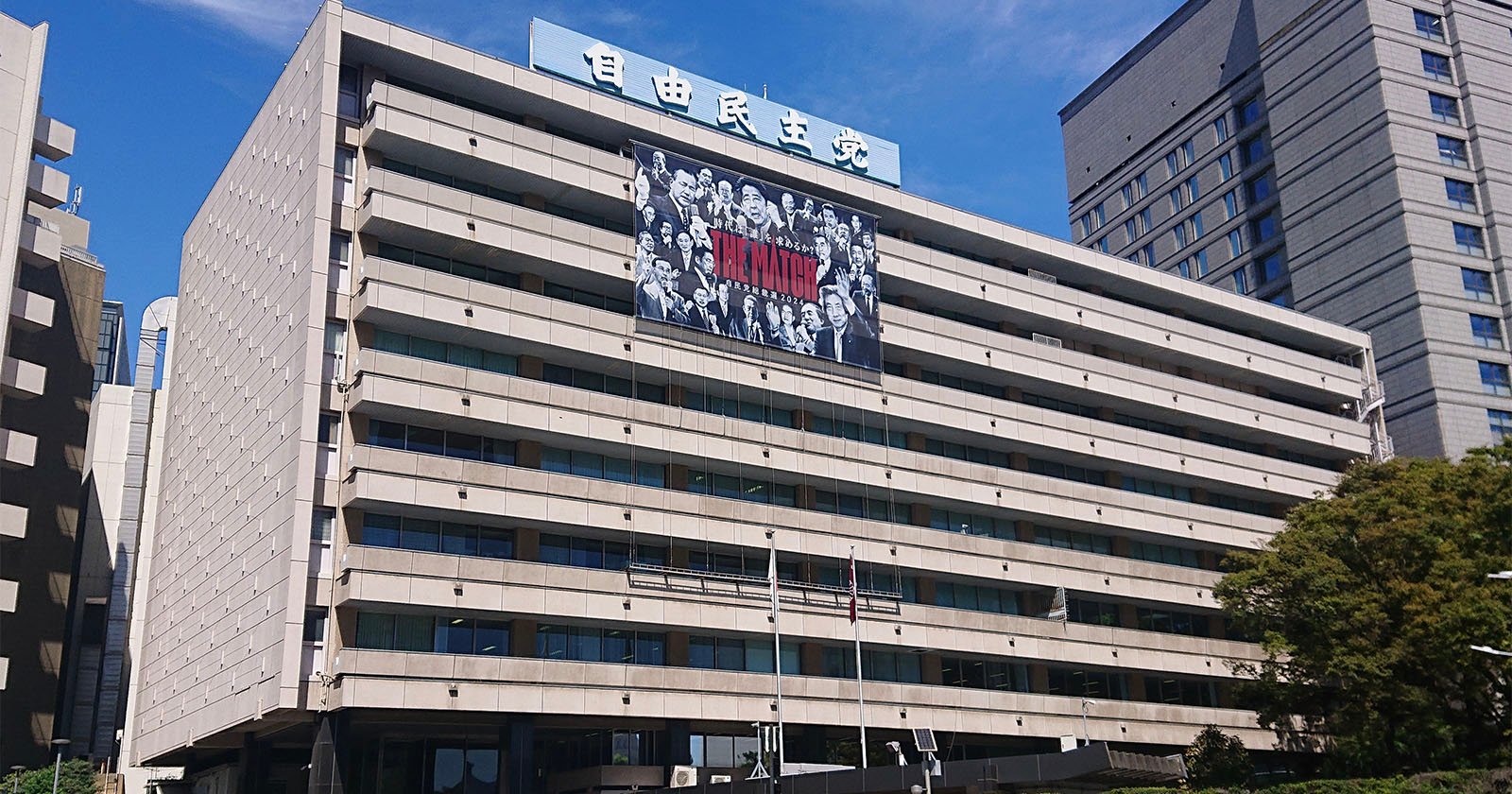 A large modern office building with a Japanese sign on the roof and a black-and-white banner featuring characters from the anime Attack on Titan displayed across its facade. Blue sky and other tall buildings are in the background. A large modern office building with a Japanese sign on the roof and a black-and-white banner featuring characters from the anime Attack on Titan displayed across its facade. Blue sky and other tall buildings are in the background.