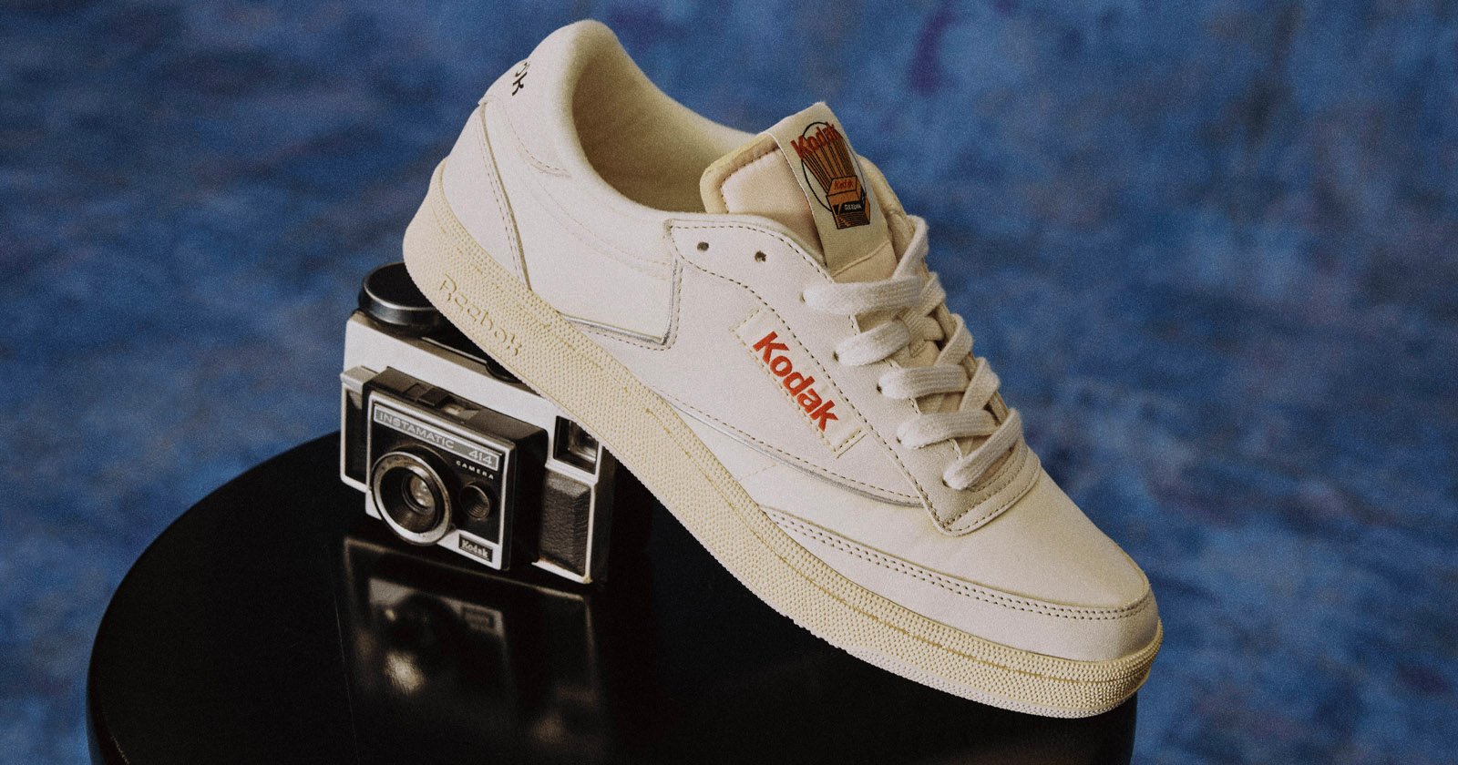 A cream-colored sneaker with Kodak branding rests on top of a vintage Kodak camera, set against a blue textured background. A cream-colored sneaker with Kodak branding rests on top of a vintage Kodak camera, set against a blue textured background.
