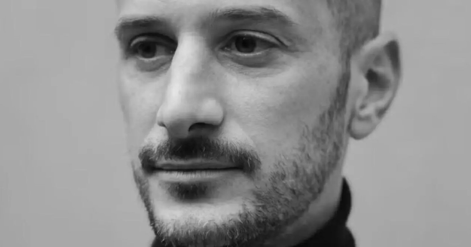 A black-and-white close-up photo of a man with a trimmed beard and mustache, wearing a dark shirt, looking slightly to the side with a neutral expression. A black-and-white close-up photo of a man with a trimmed beard and mustache, wearing a dark shirt, looking slightly to the side with a neutral expression.