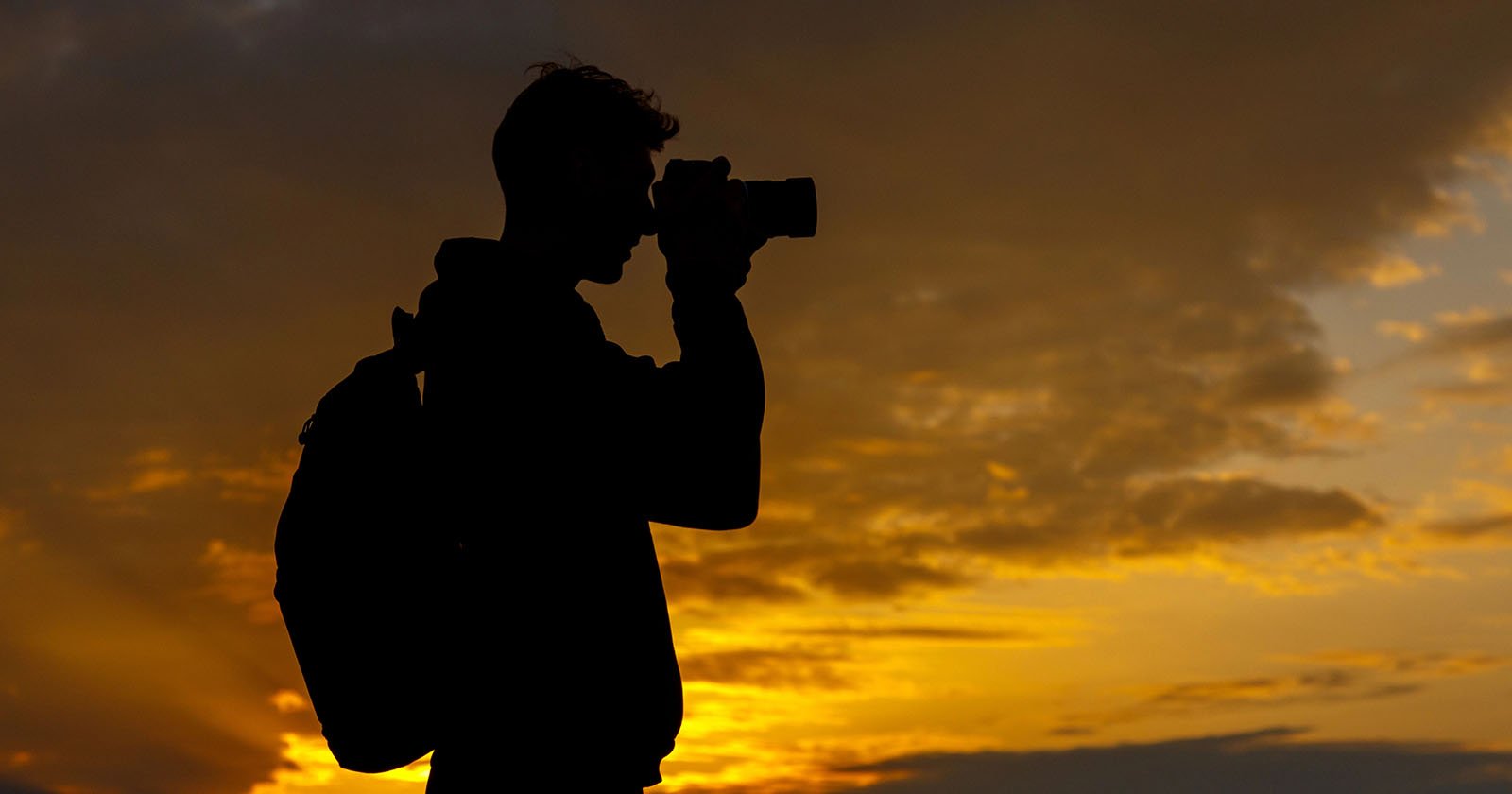 Silhouette of a person with a backpack taking a photo with a camera against a dramatic, golden sunset sky with scattered clouds. Silhouette of a person with a backpack taking a photo with a camera against a dramatic, golden sunset sky with scattered clouds.