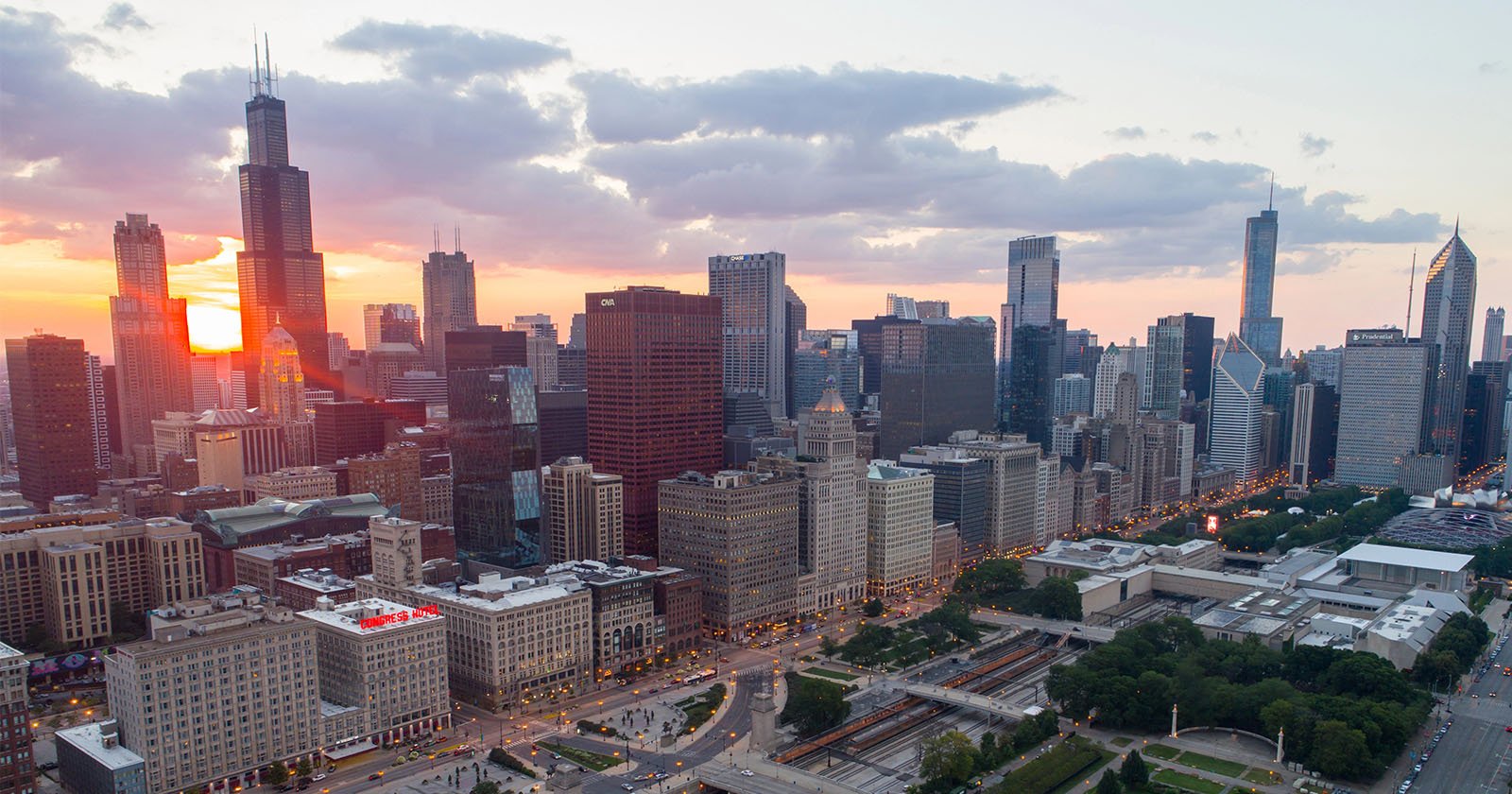 Aerial view of a city skyline at sunset, with sunlight reflecting off tall skyscrapers and clouds in the sky. Streets and green spaces are visible in the foreground. Aerial view of a city skyline at sunset, with sunlight reflecting off tall skyscrapers and clouds in the sky. Streets and green spaces are visible in the foreground.